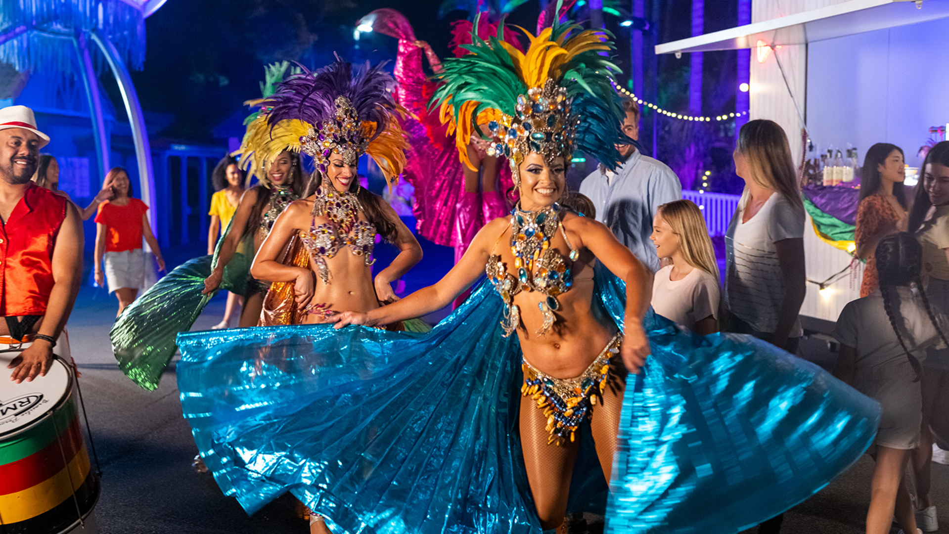 Two women in colorful feathered headdresses and sparkling costumes dance with blue capes at a lively outdoor festival at night, surrounded by people, lights, and a drummer.