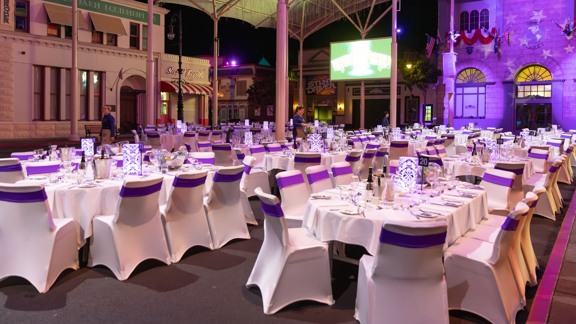 Rows of round tables with white tablecloths and chairs covered in white fabric with purple sashes are set for an outdoor event at night, surrounded by decorative buildings and illuminated by purple lighting.