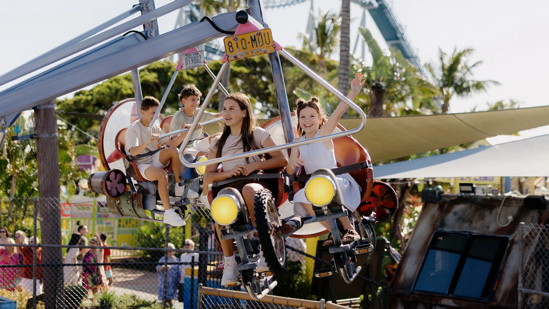 Four people ride a suspended amusement park ride shaped like a car with bike pedals. Palm trees and other park attractions are visible in the background. One child is waving and smiling excitedly.