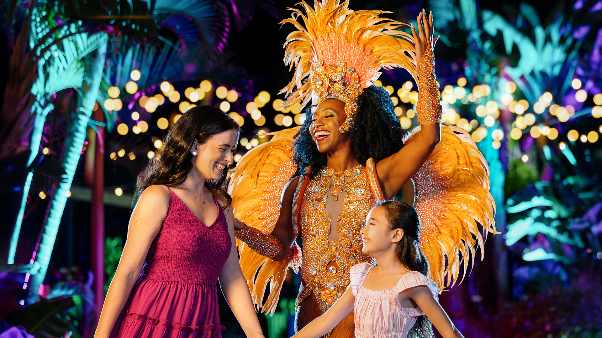 A vibrant performer in an elaborate orange feathered costume greets a smiling woman and young girl at night, surrounded by colorful lights and tropical plants.