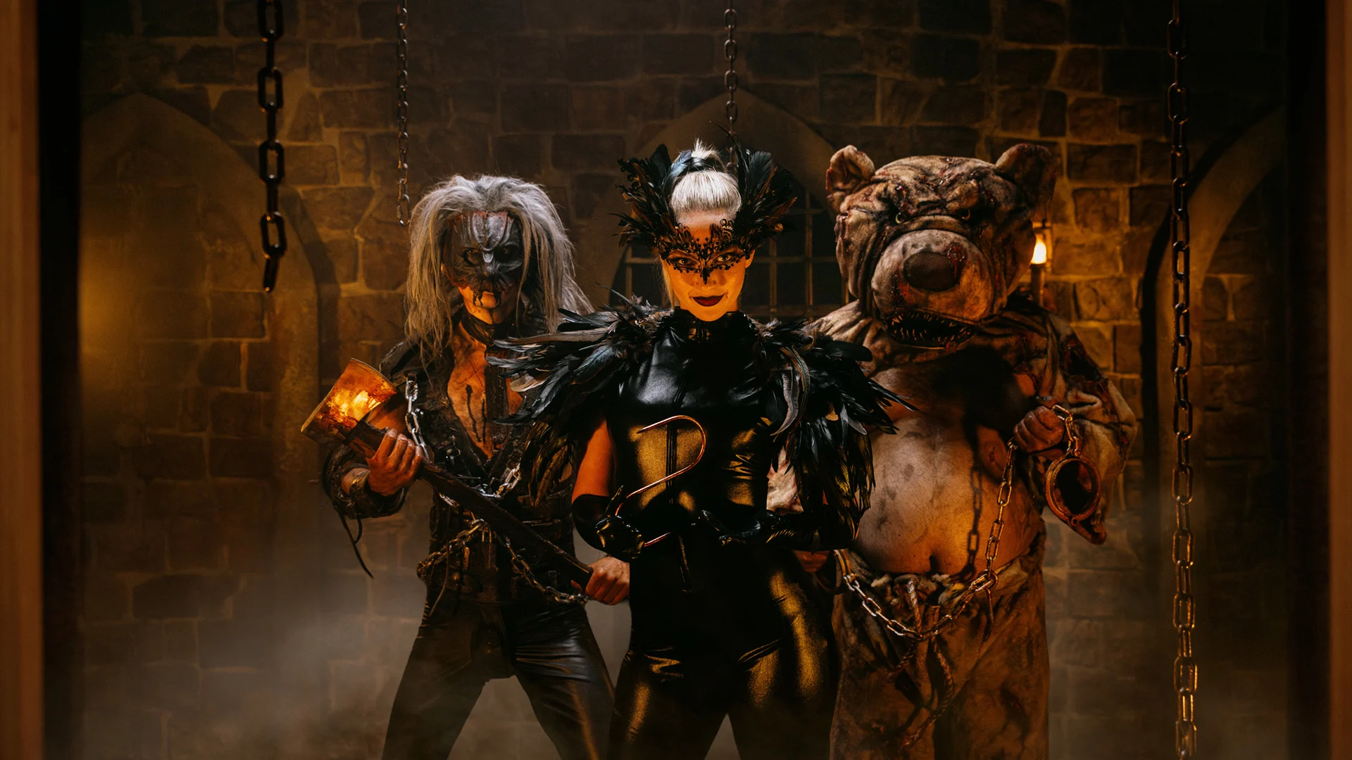 Three people in elaborate costumes stand in a dimly lit stone room. One wears a beastly mask and holds an axe, another is in black with a feathered mask, and the third is in a bear suit holding a torch. Chains hang around them.
