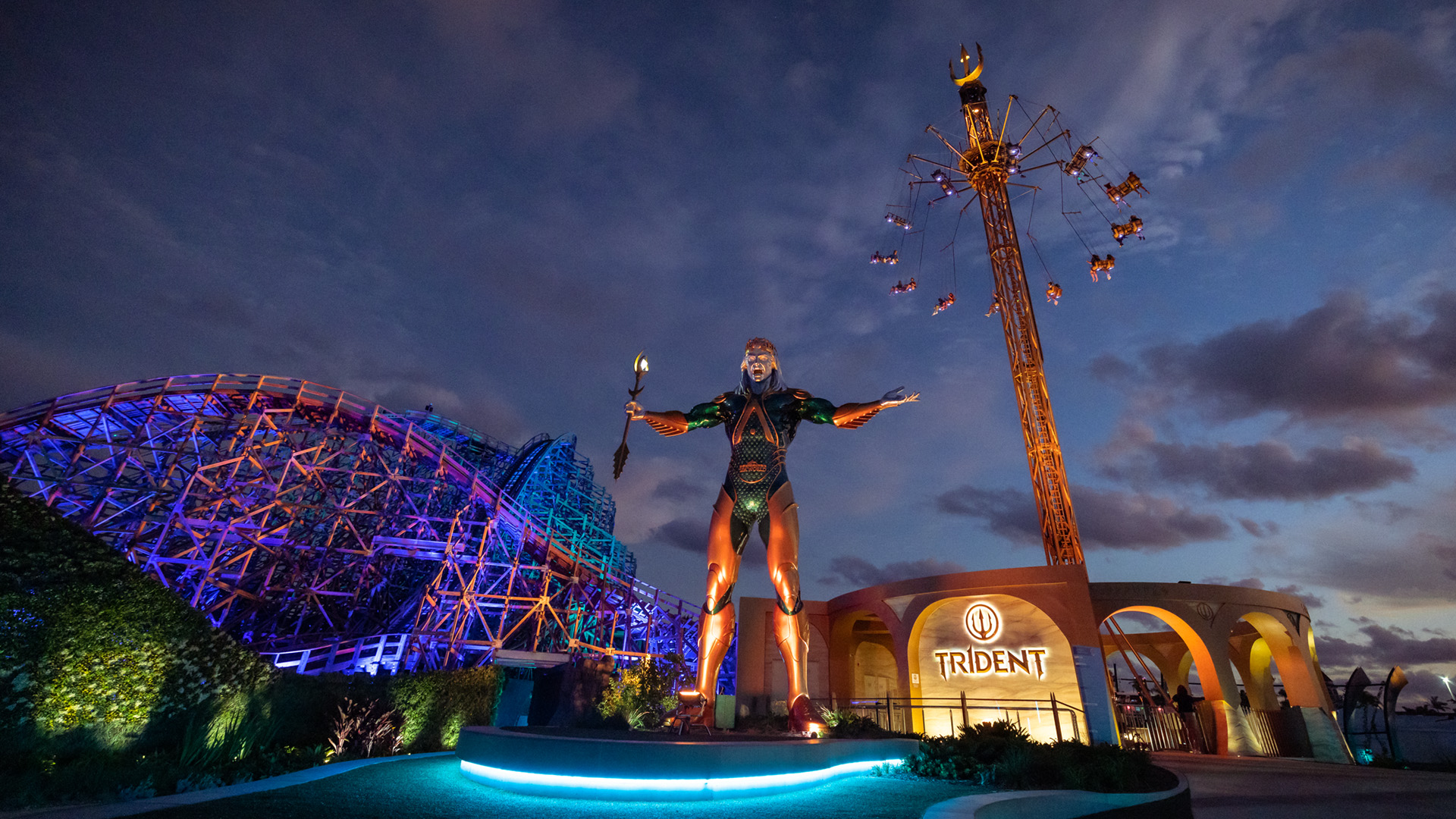 A large, illuminated trident-wielding statue stands in front of a roller coaster and a tall spinning ride at an amusement park during dusk, with vibrant lights and a dramatic, cloudy sky.
