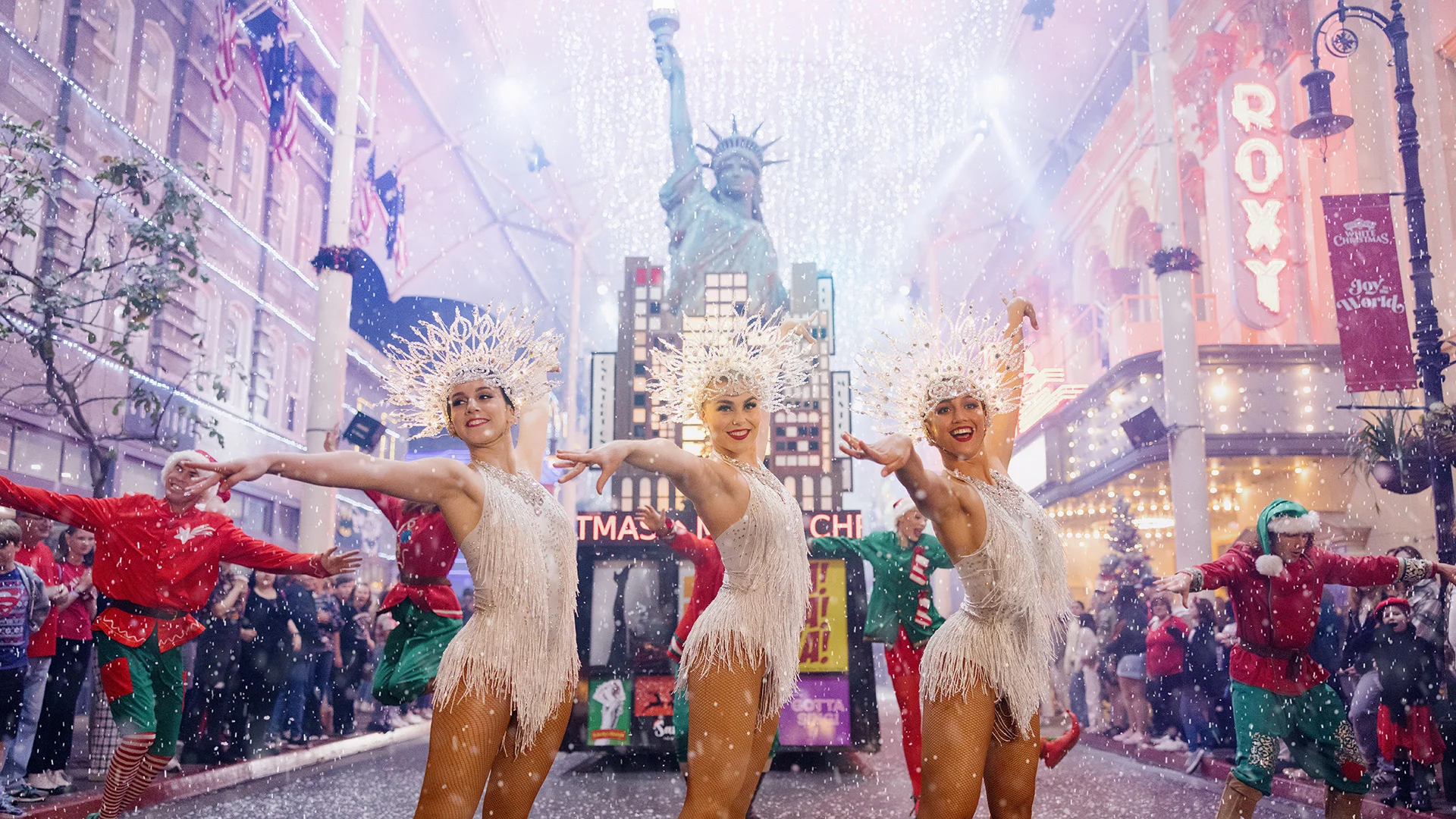 Three dancers in white fringed costumes and sparkling headdresses perform in falling snow on a festive city street, with the Statue of Liberty replica and bright holiday lights in the background.