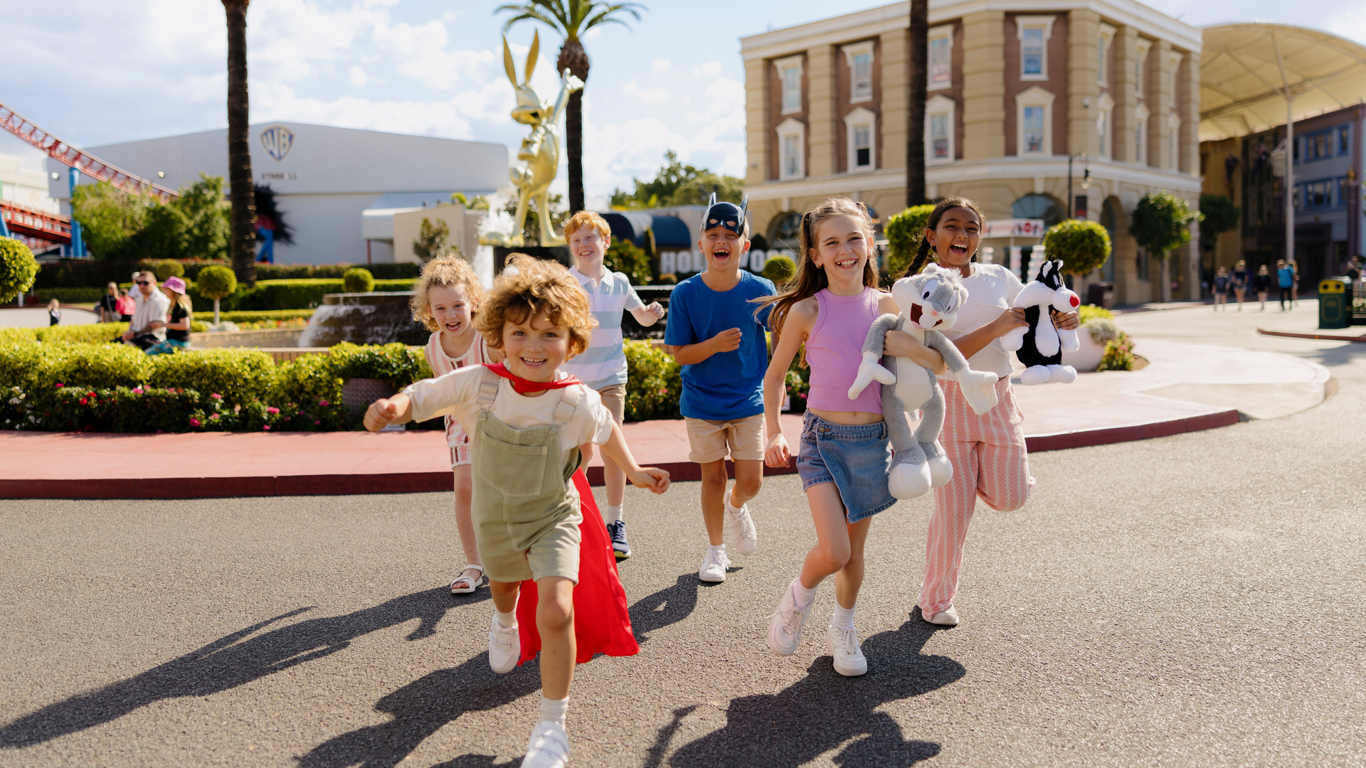 A group of smiling children run toward the camera on a sunny day at an amusement park, carrying plush toys and wearing casual clothes, with buildings and a fountain in the background.