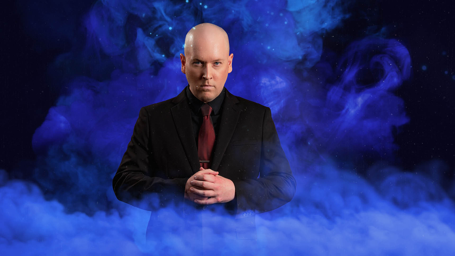 Bald man in a black suit and red tie stands with hands clasped, in front of a swirling green smoke background.