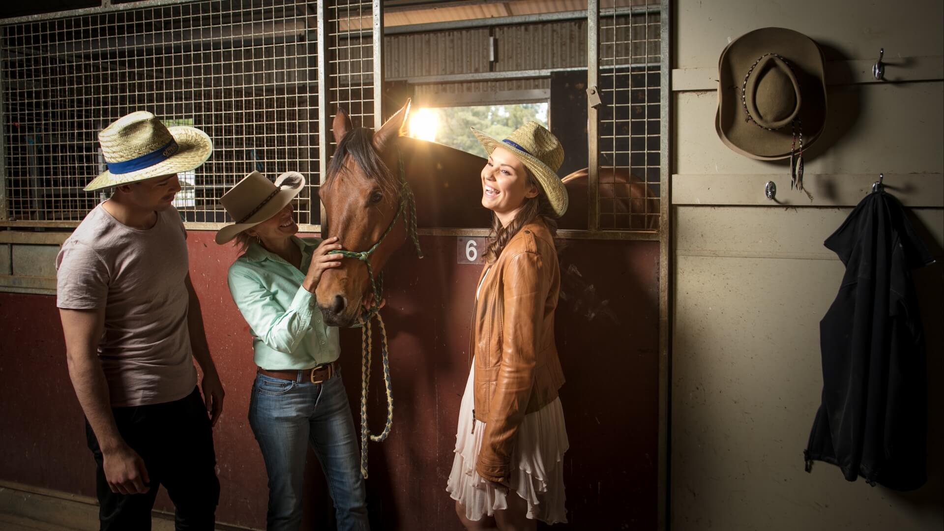 Three people in cowboy hats stand beside a horse in a stable. They are smiling and interacting with the horse.