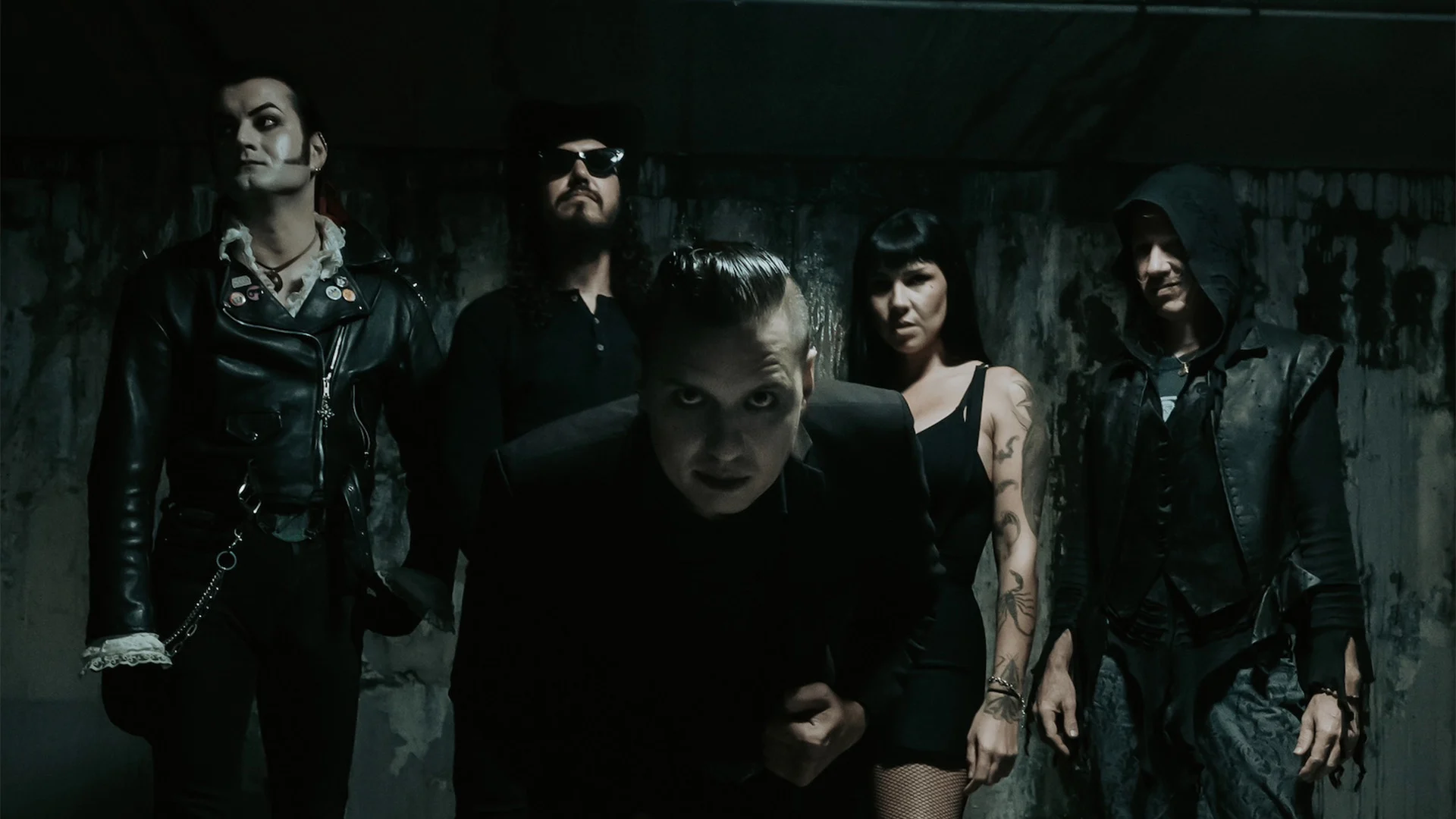 Five people in dark, edgy clothing stand in a dimly lit, grungy room. The person in front leans toward the camera, while the others pose behind, creating a mysterious and intense atmosphere.