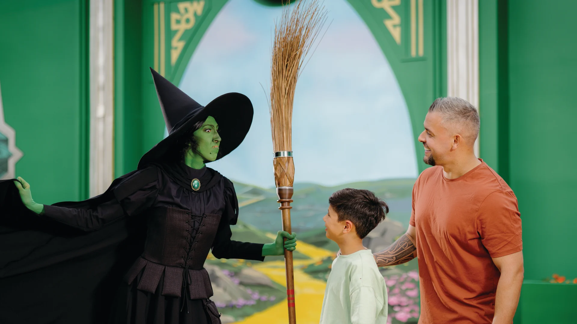 A person dressed as the Wicked Witch of the West, holding a broom, stands next to a smiling man and boy in front of a yellow brick road backdrop from "The Wizard of Oz.