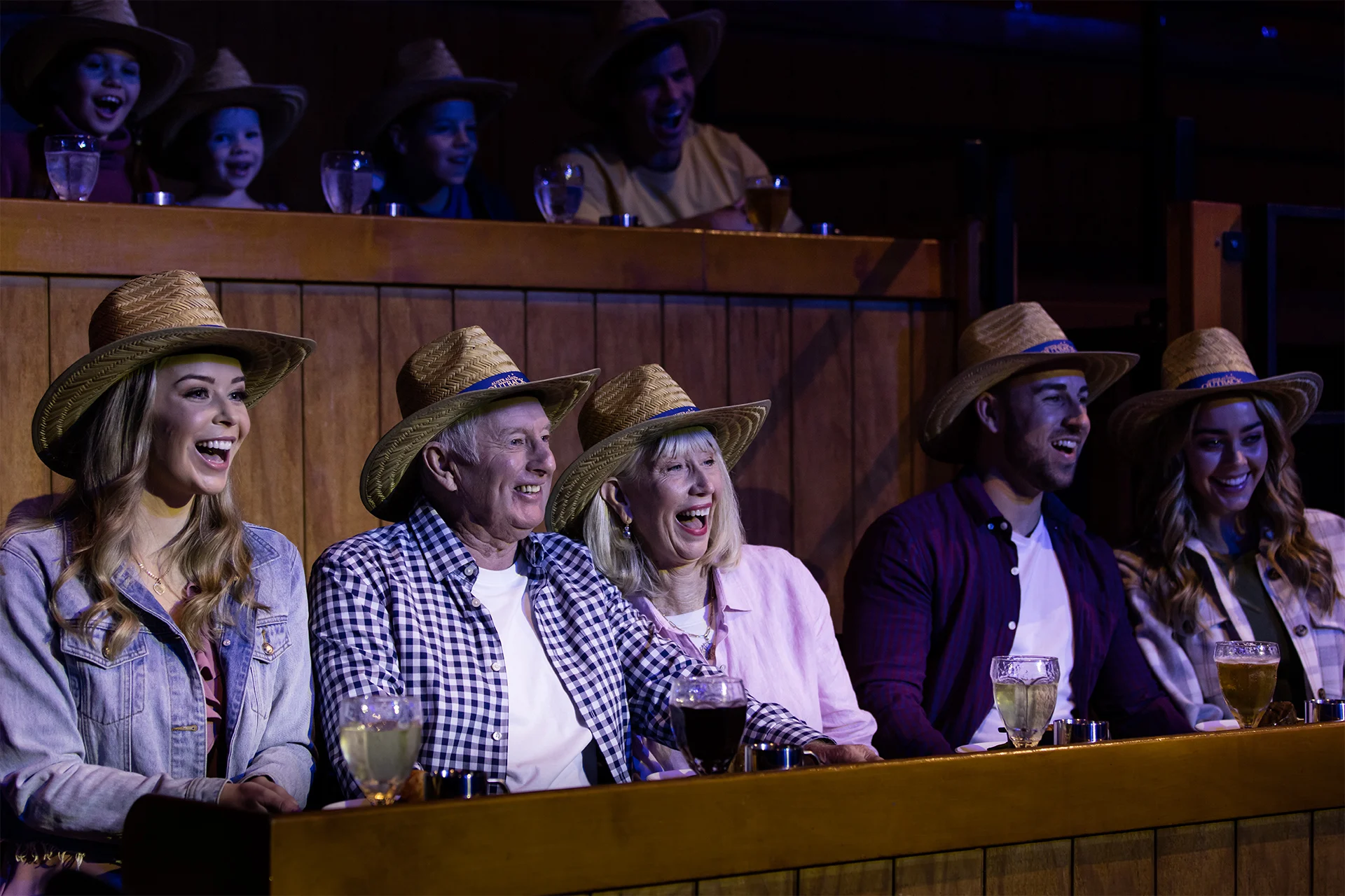 A group of people wearing straw hats sit in an audience, laughing and watching a performance. Drinks rest on the railing in front of them.