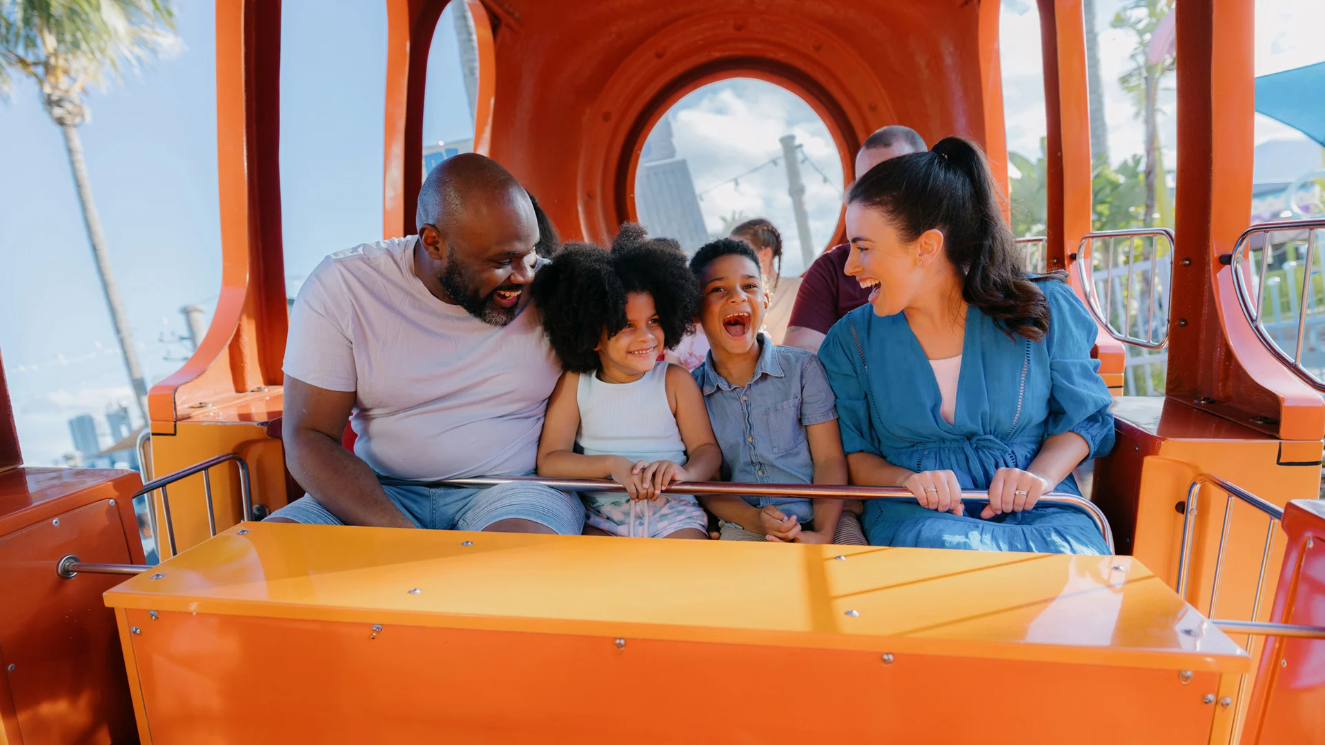 A family of four smiles and laughs while riding a theme park ride at Sea World. They are seated in an orange and red carriage with other passengers visible in the background.