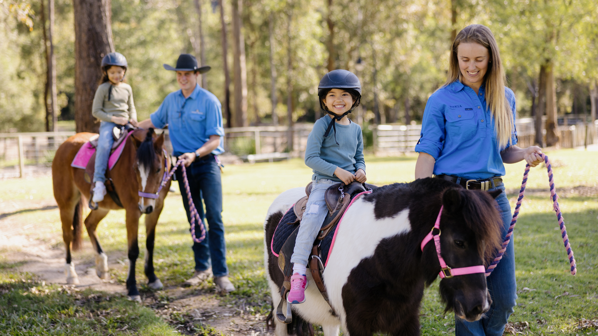 Two children wearing helmets ride ponies, each led by an adult on a grassy path in a wooded area. Both adults are dressed in blue shirts, and the children appear happy and engaged.