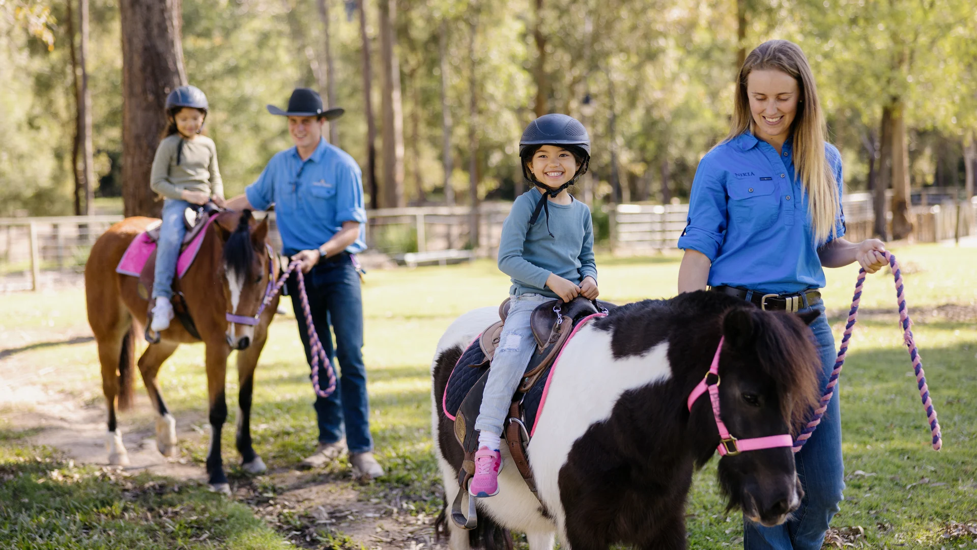 Two children wearing helmets ride ponies, each led by an adult on a grassy path in a wooded area. Both adults are dressed in blue shirts, and the children appear happy and engaged.