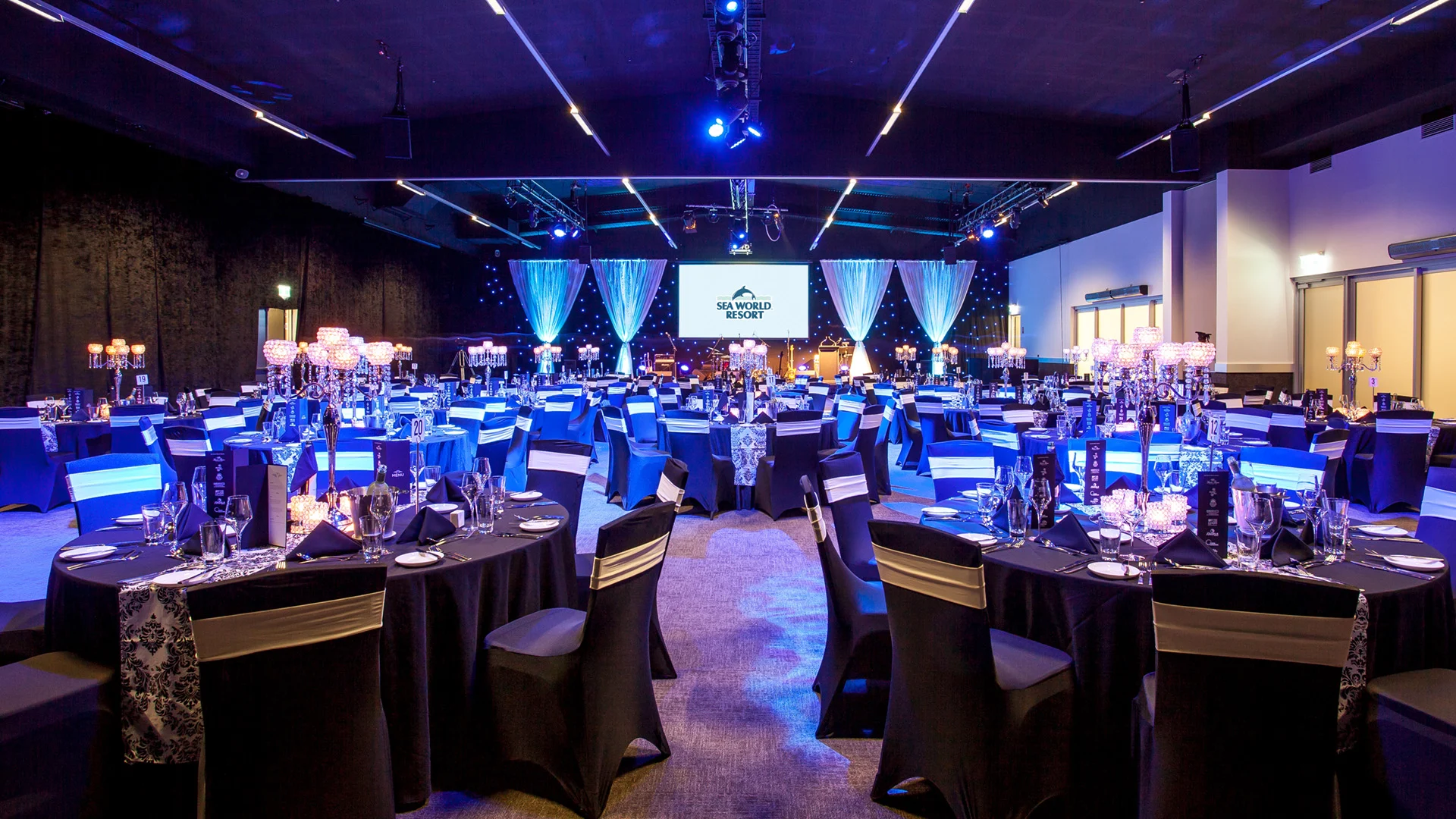 A spacious event hall with round tables covered in black and white cloths, set for a formal occasion. Blue lighting illuminates the room, and a large screen is visible at the front.