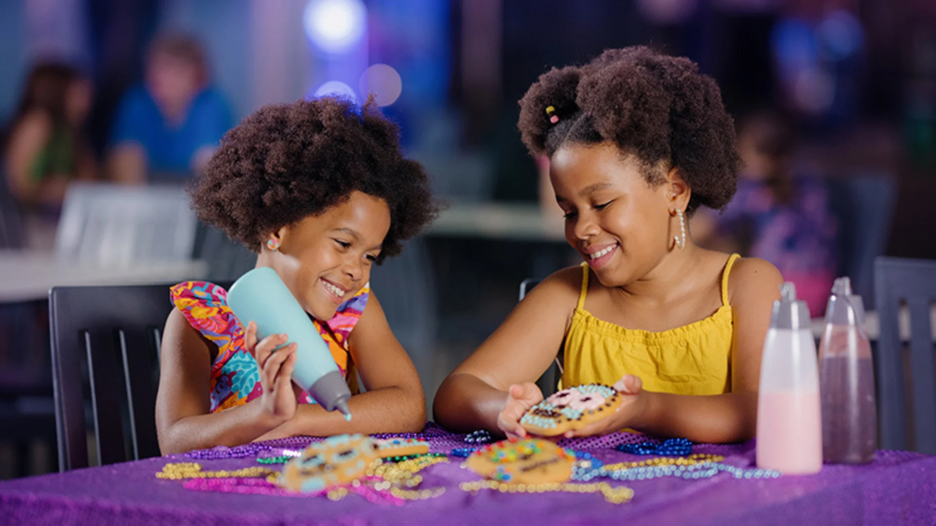 Two young girls with curly hair sit at a table decorated with beads, smiling as they decorate cookies with colorful icing at what appears to be a fun event or party.