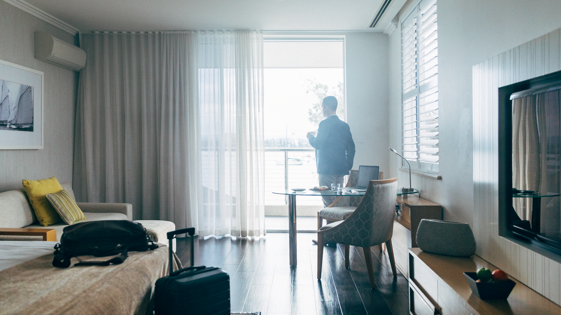 A person stands by a window in a modern hotel room with a suitcase, armchair, table with laptop, and a wall-mounted TV.