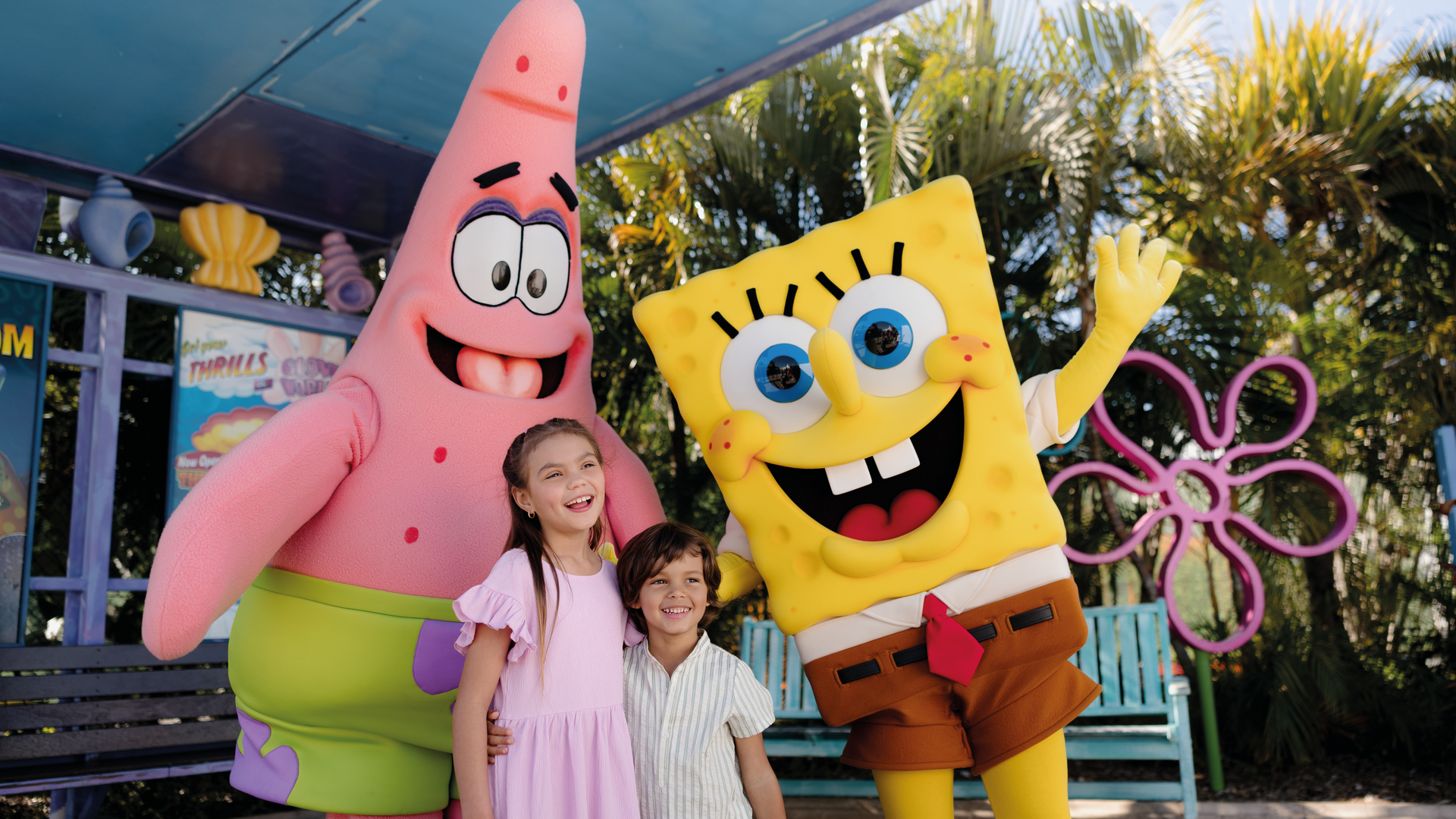 Two children smile and pose with people in large Patrick Star and SpongeBob SquarePants costumes outdoors, surrounded by palm trees and colorful decorations.