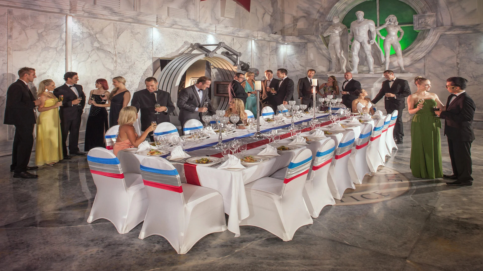 A group of elegantly dressed people stand and sit around a long banquet table with white chairs and tablecloths in a grand, marble hall decorated with large statues.