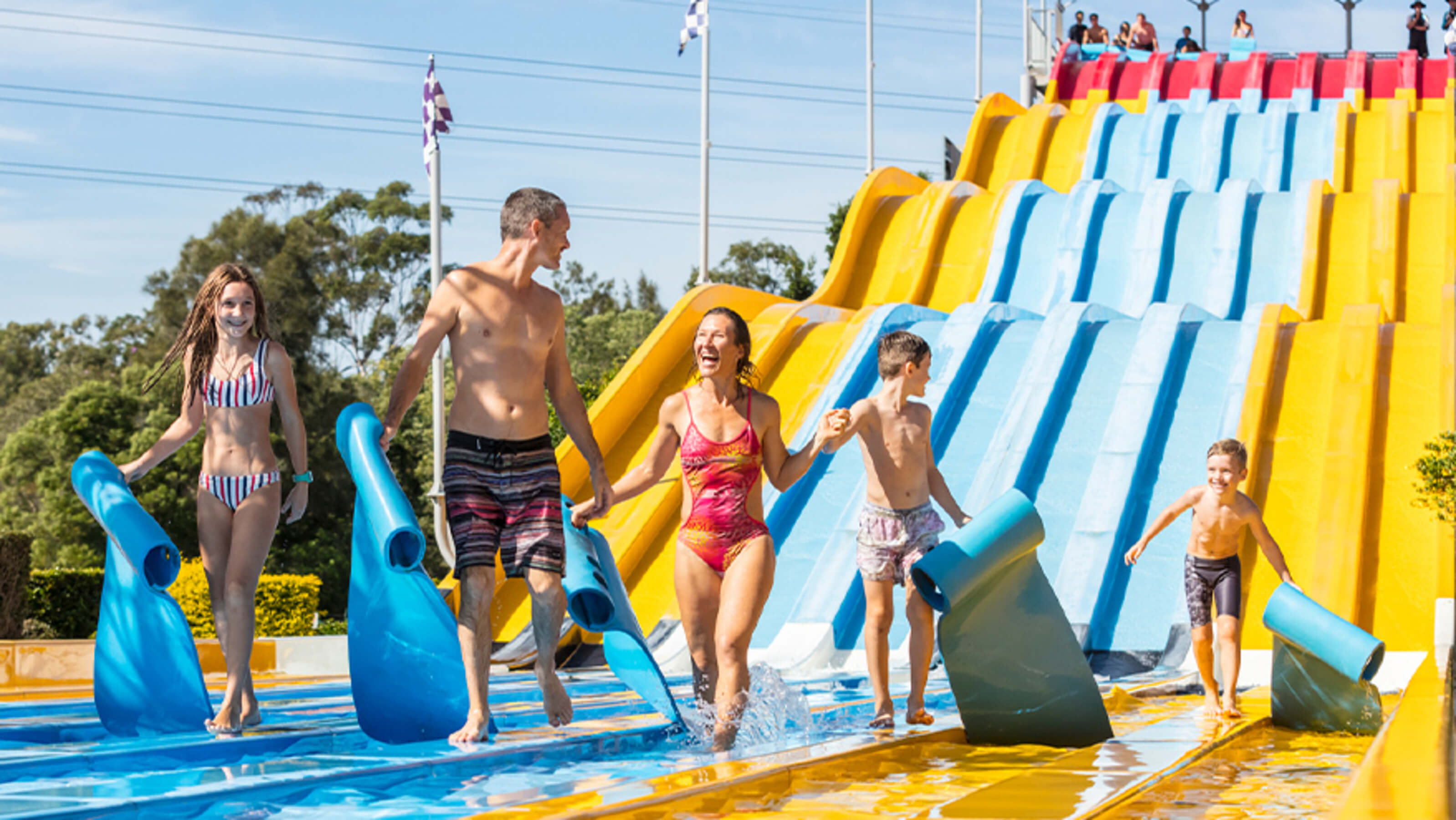 Family laughing and holding mats after racing down the colourful multi-lane Aqua Racer water slide at Wet’n’Wild Gold Coast on a sunny day