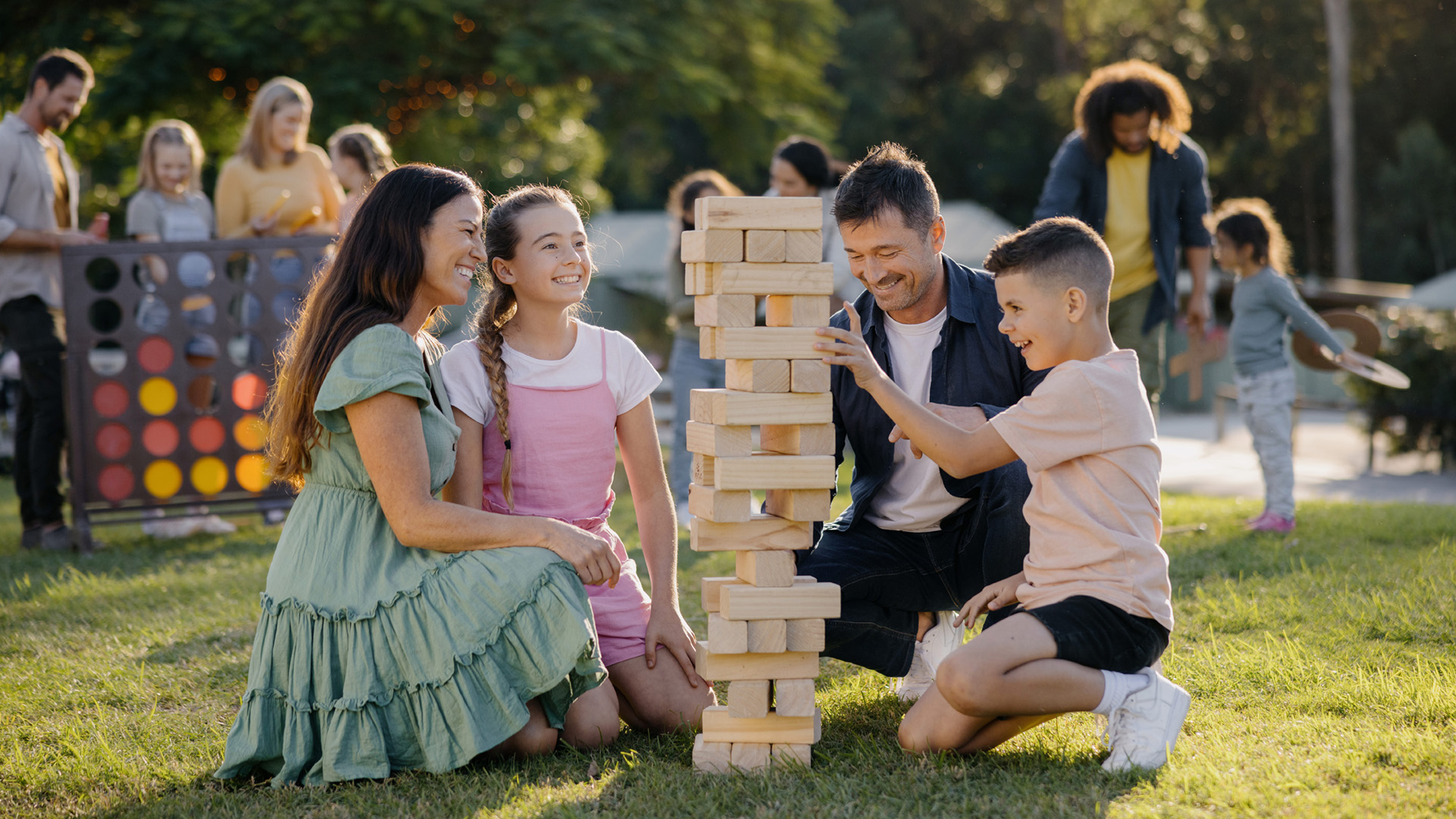 A family of four kneels on grass, playing a large outdoor Jenga game, while other people enjoy activities in the background at a park.