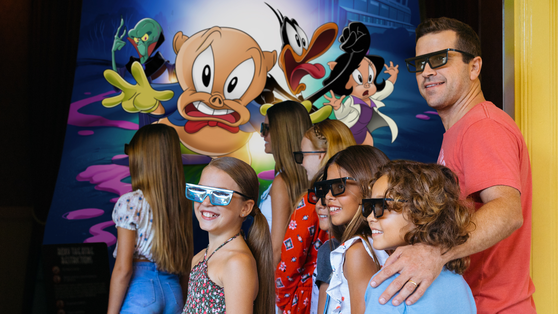 A group of children and an adult wearing 3D glasses smile in front of a colorful cartoon display featuring Looney Tunes characters.