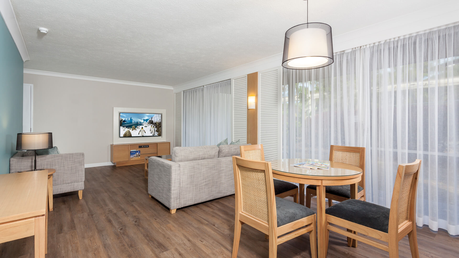 Modern living room and dining area with light wood flooring, gray sofas, round dining table with four chairs, flat-screen TV, and large windows with sheer curtains.