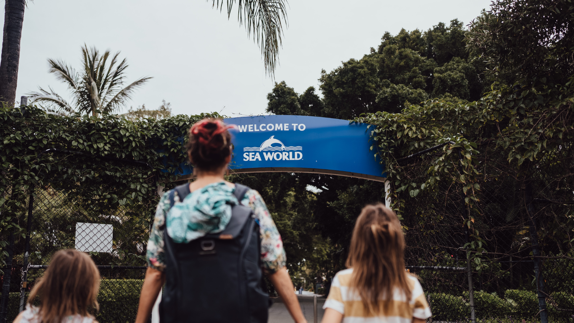 A woman and two children walk toward a blue "Welcome to SeaWorld" sign at the entrance, surrounded by greenery and trees.