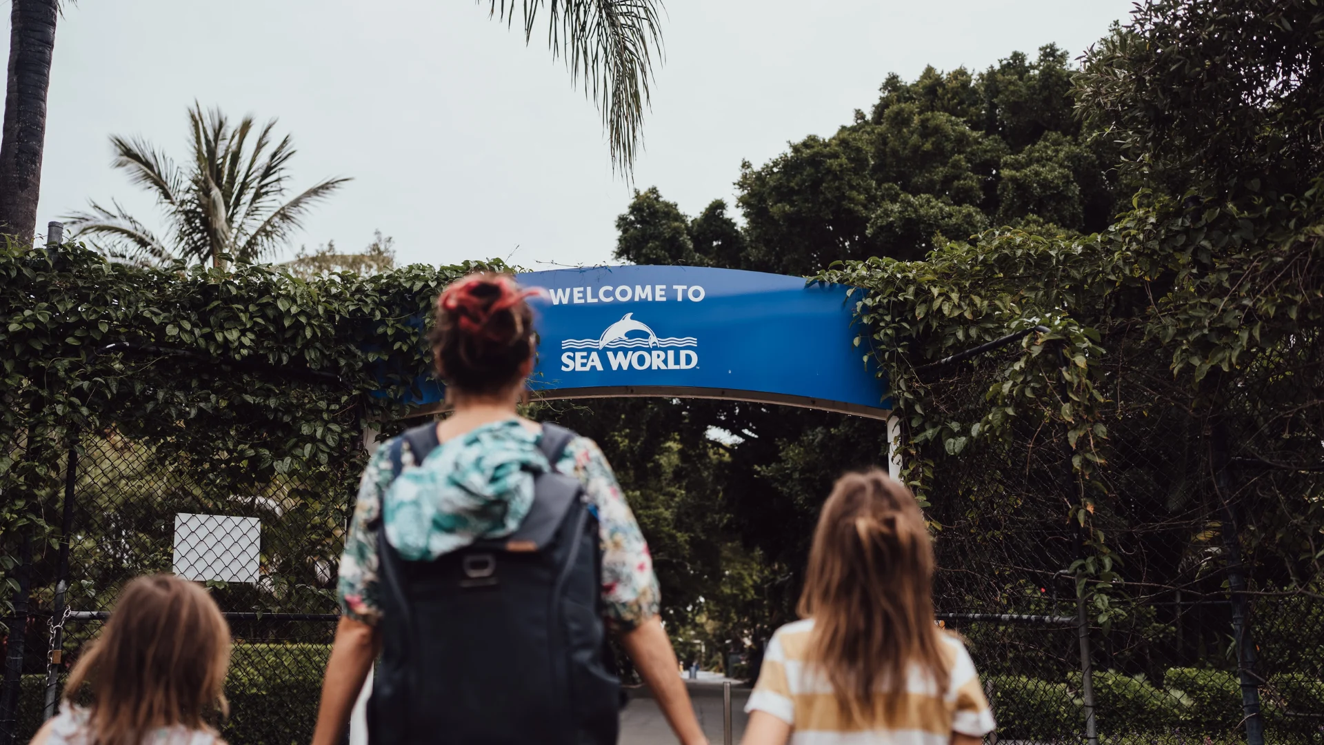 A woman and two children walk toward a blue "Welcome to SeaWorld" sign at the entrance, surrounded by greenery and trees.