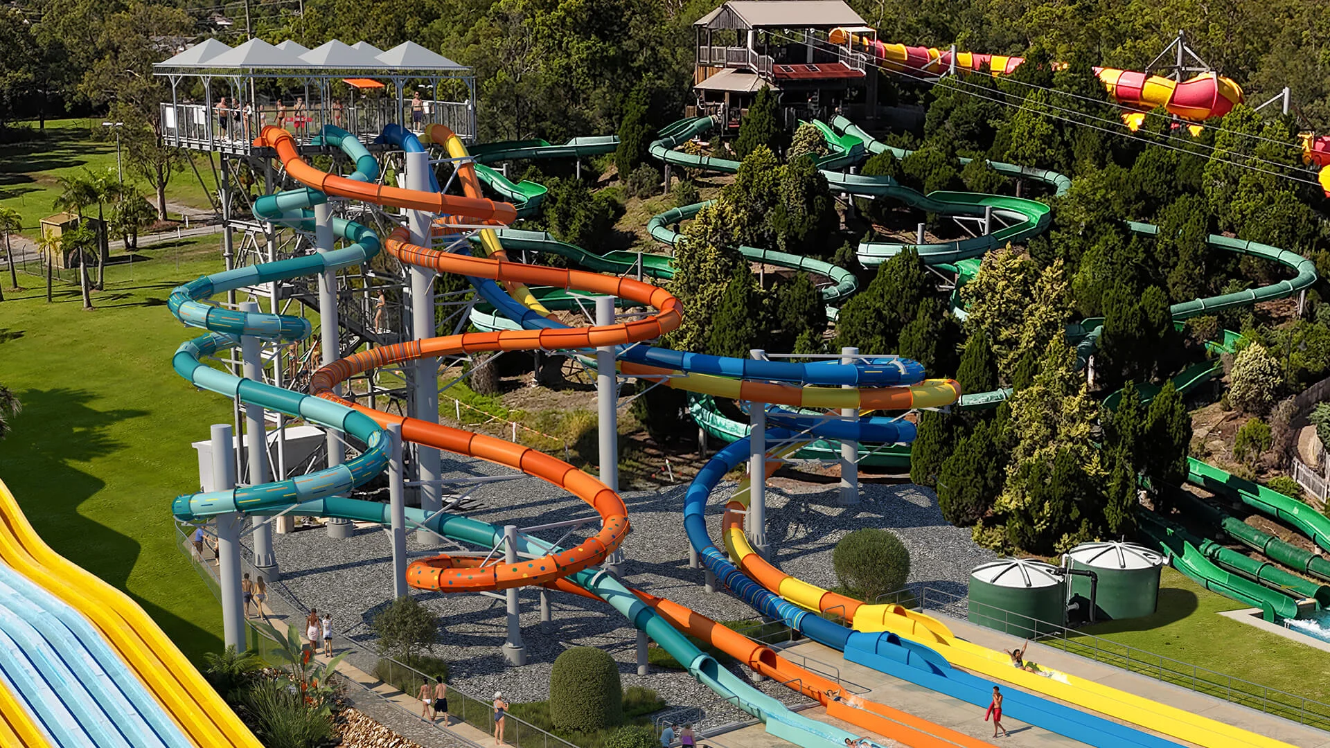 A water park features multiple twisting water slides in blue, orange, and green, surrounded by trees and grassy areas, with people walking nearby.