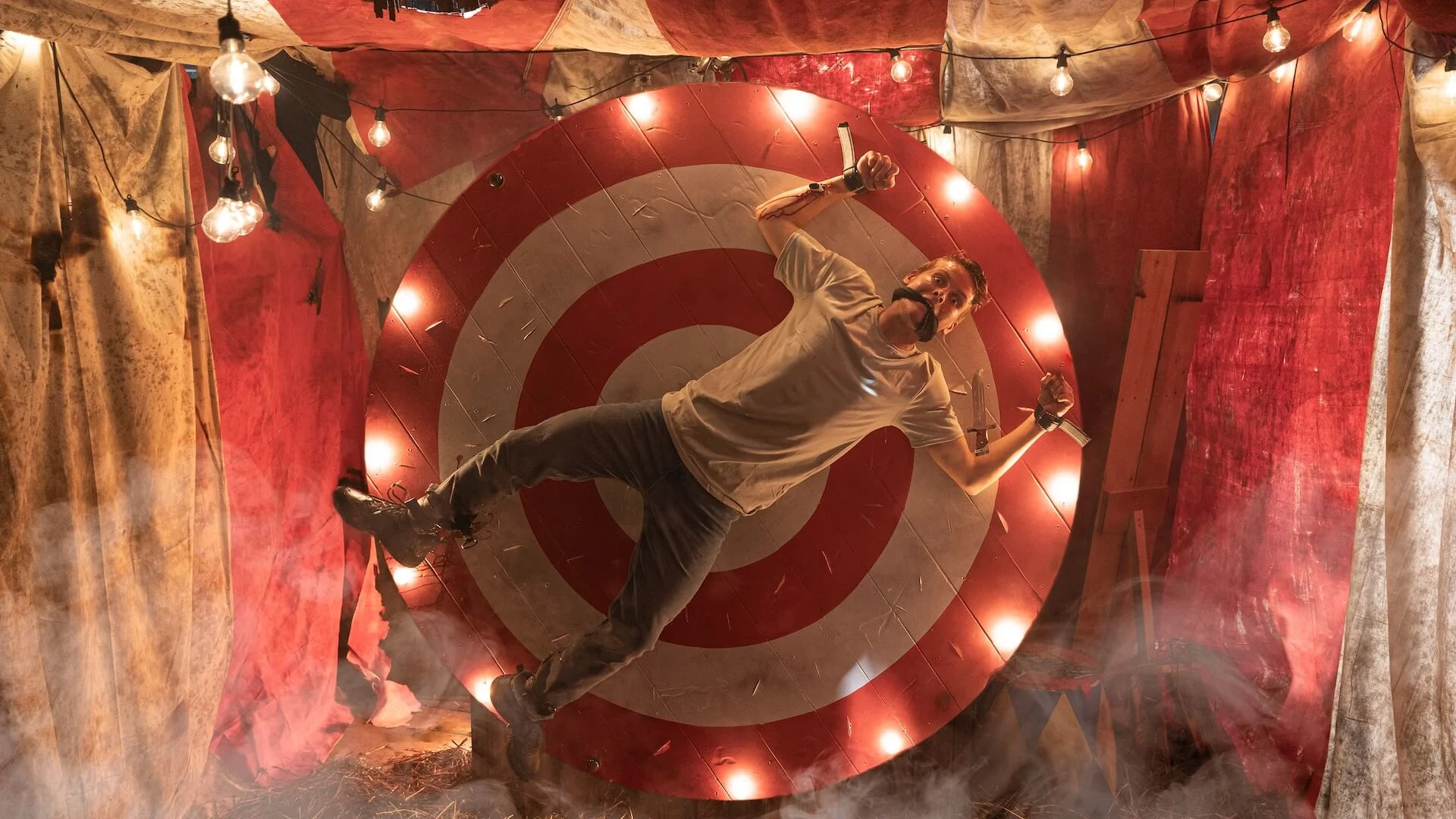 Fright Nights night event at Warner Bros Movie World. A person is strapped to a rotating red and white target wheel in a carnival setting, surrounded by red drapery and string lights.