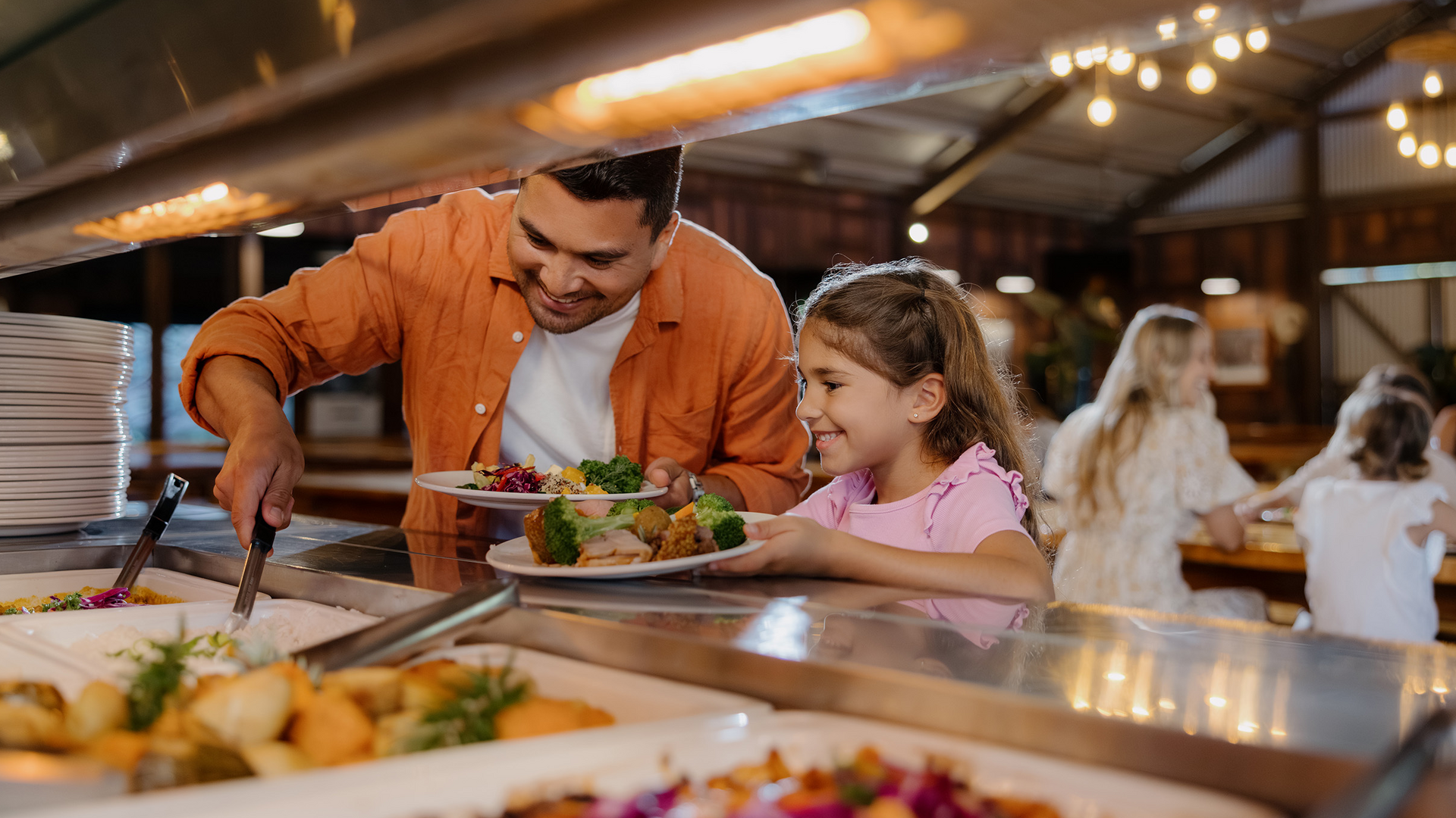 A man and a young girl are smiling as they serve themselves food from a buffet. The man is using tongs to add food to his plate while the girl holds her plate and watches. Other people dine in the background.