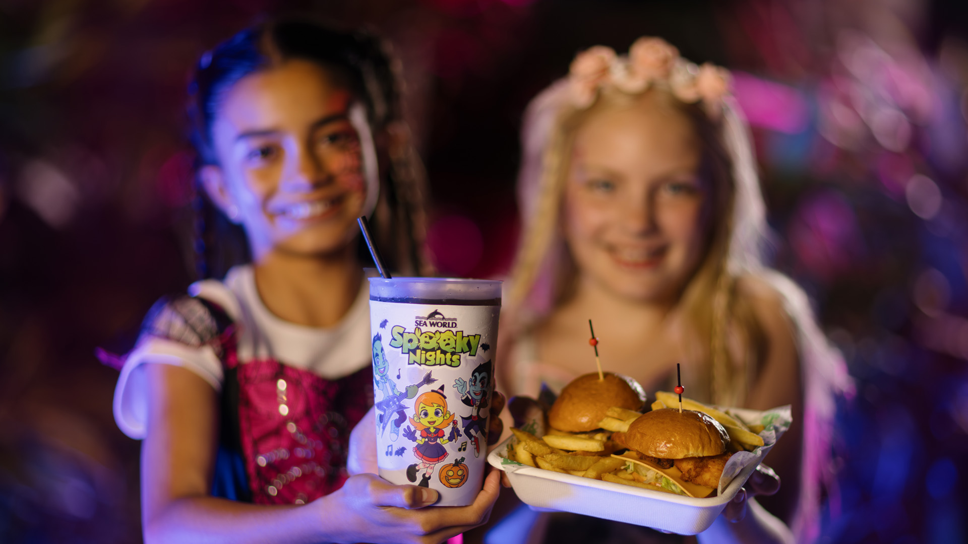 Two smiling children in costumes hold food—one with a Halloween-themed drink cup, the other with a tray of sliders and fries—against a colorful, blurred background.