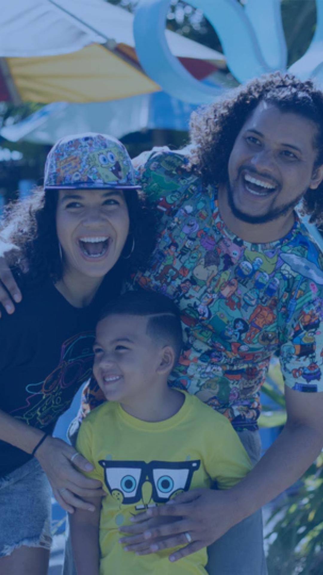 Three people, two adults and one child, stand close together outdoors, smiling widely. The adults wear colorful shirts, and the child wears a yellow T-shirt.