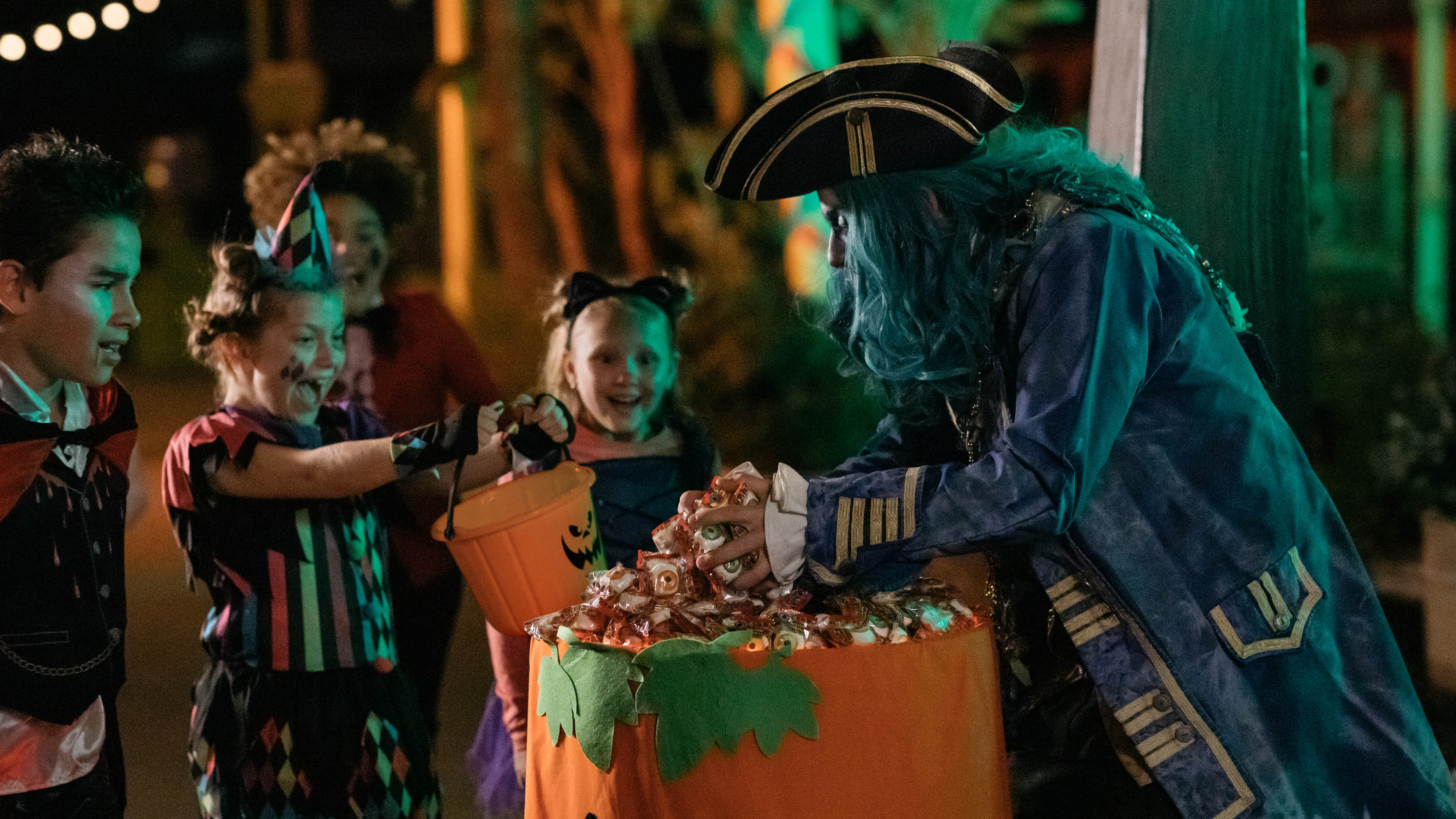Children in Halloween costumes smile and hold candy buckets while a person dressed as a pirate with blue hair hands out candy from a large container outdoors at night.