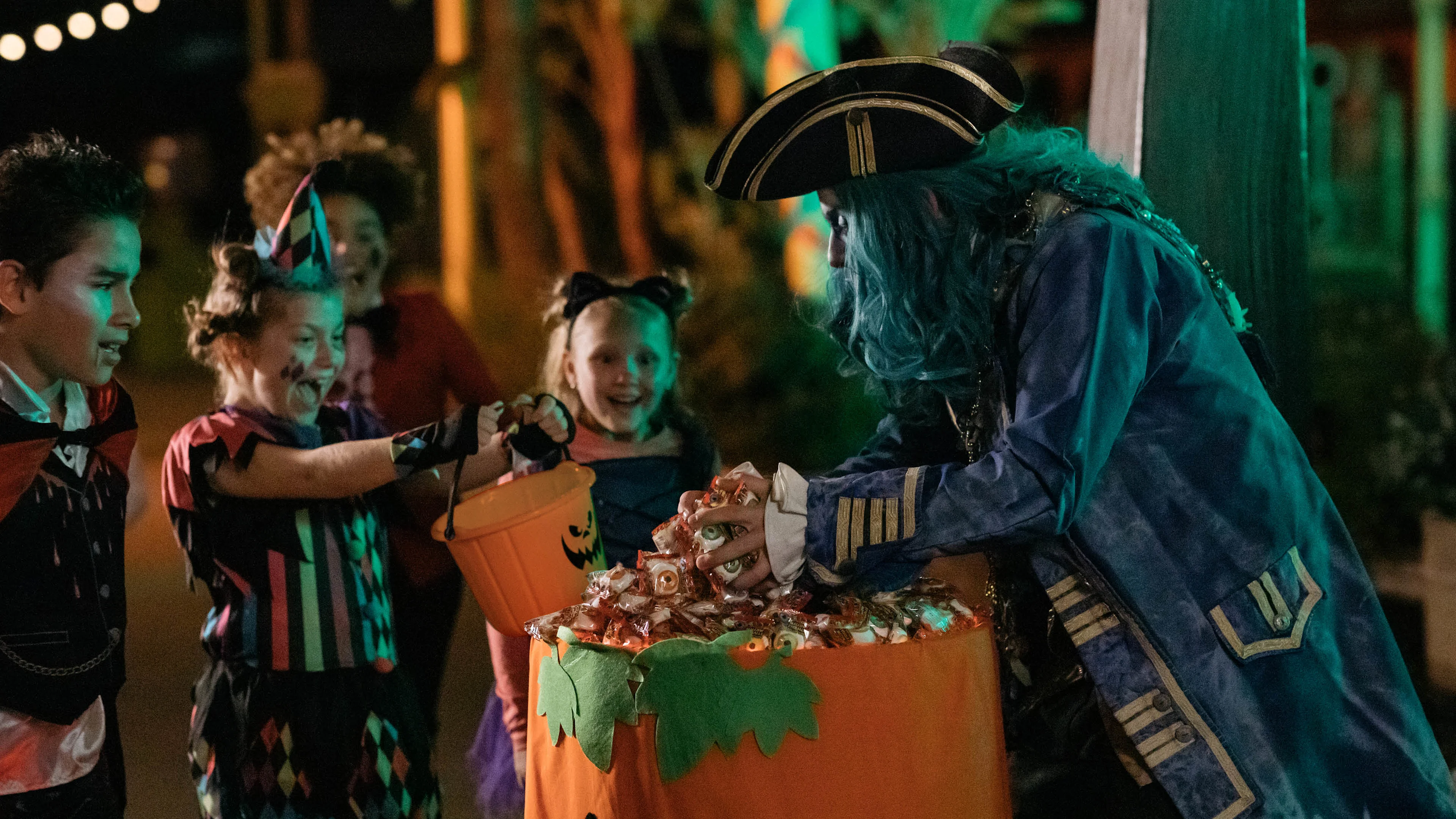 Children in Halloween costumes smile and hold candy buckets while a person dressed as a pirate with blue hair hands out candy from a large container outdoors at night.