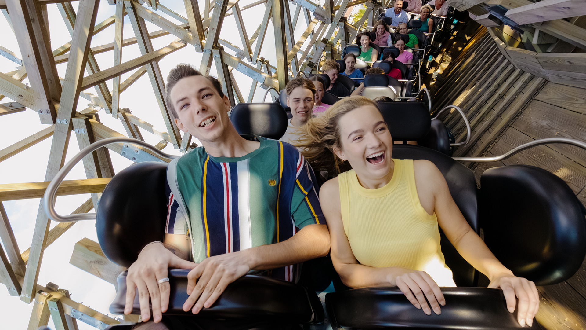 Two young adults, a man and a woman, smile and laugh while riding a wooden roller coaster with other excited riders behind them under a bright, sunny sky.