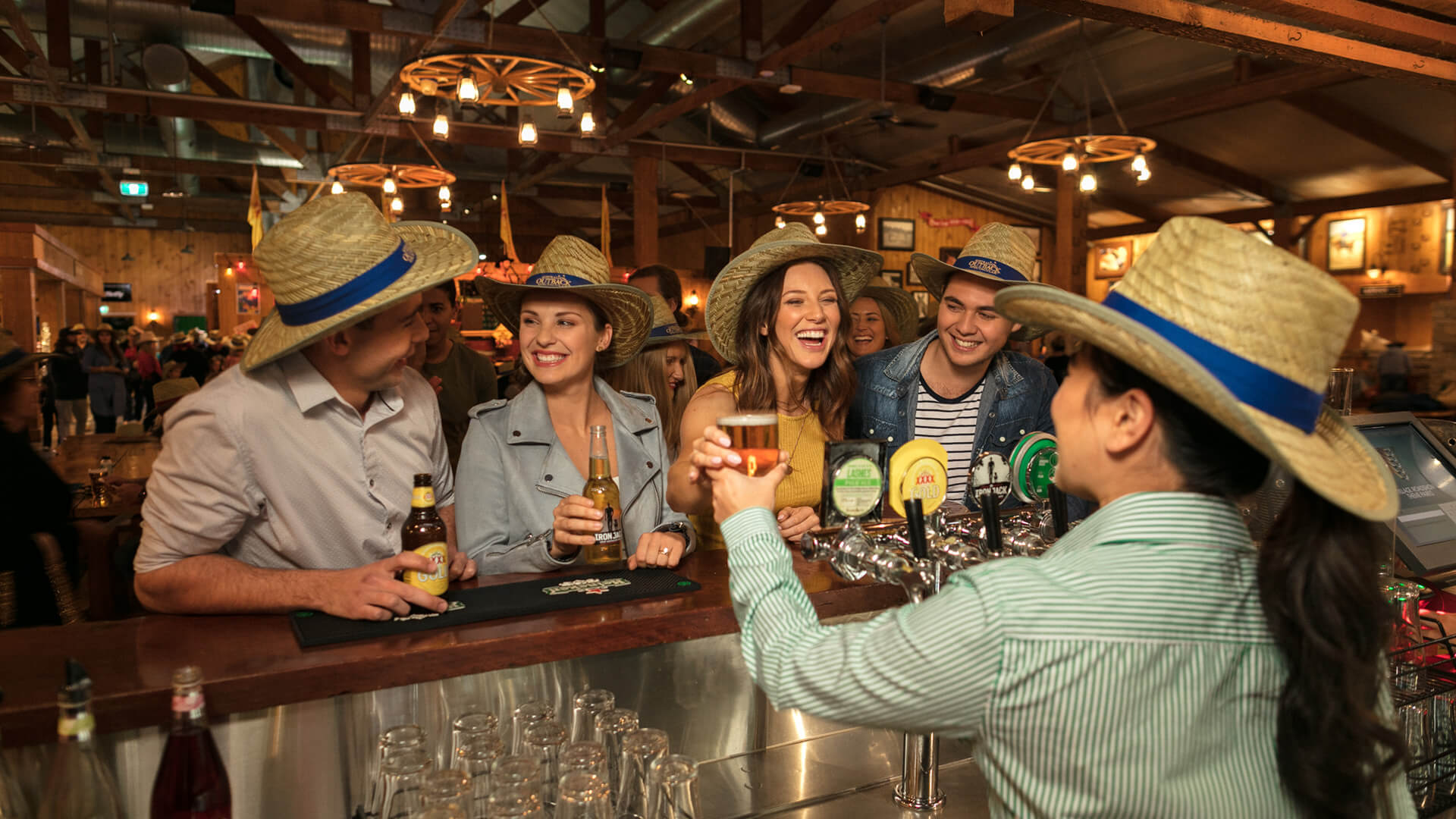 A group of people wearing straw hats are standing at a bar at Australian Outback Spectacular, engaging in conversation with a bartender who is serving drinks. The setting appears lively and rustic.