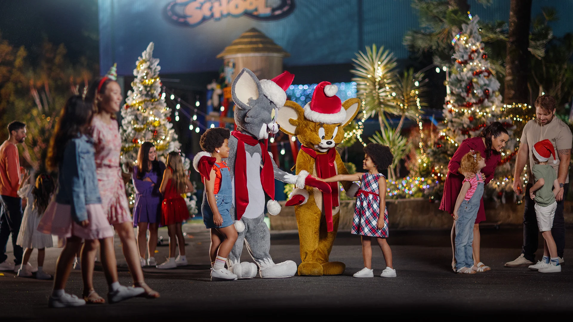 Children and adults gather outdoors at night, interacting with people in Tom and Jerry costumes wearing Santa hats. Christmas trees with lights and ornaments are in the background, creating a festive atmosphere.