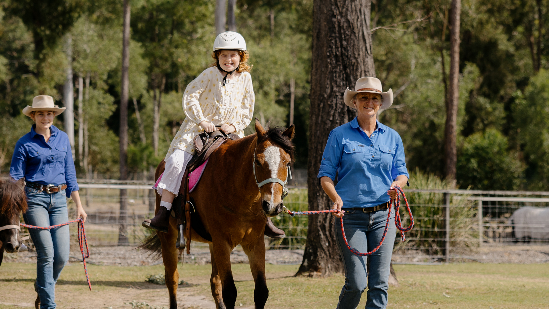 A young girl in a white helmet and dress rides a brown horse, led by a woman in a blue shirt and cowboy hat. Another woman in a blue shirt and hat walks beside the horse.