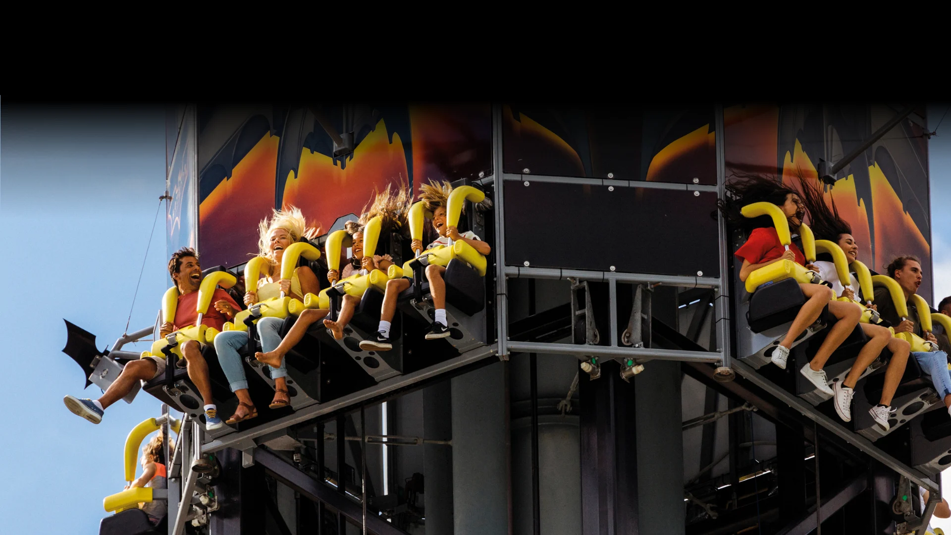 People are seated on a thrill ride, gripping safety bars, as they prepare for a drop. The background features dramatic flames and a clear blue sky above. The ride has yellow seats and black supports.