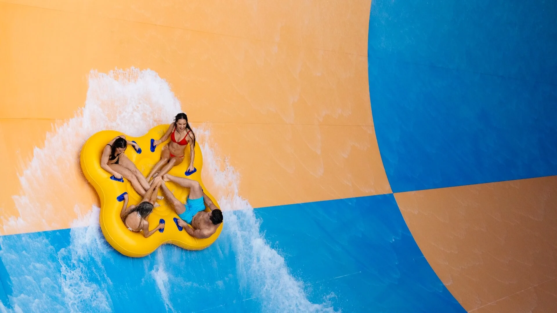 Group of friends riding a large yellow raft on Wet’n’Wild’s Tornado water slide, splashing through steep blue and orange slide walls.