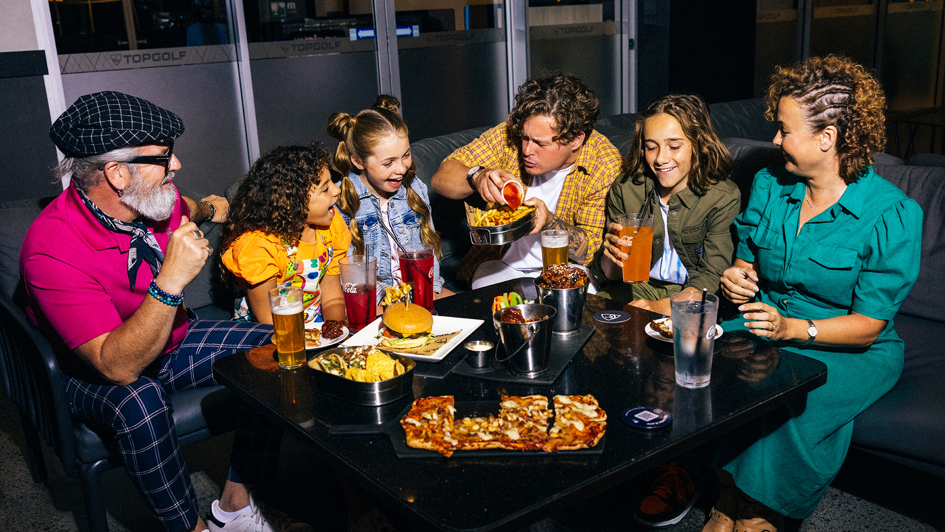 A group of six people, including adults and children, are sitting around a table at a restaurant, enjoying drinks and a variety of food like pizza and fries, laughing and sharing a lively meal together.