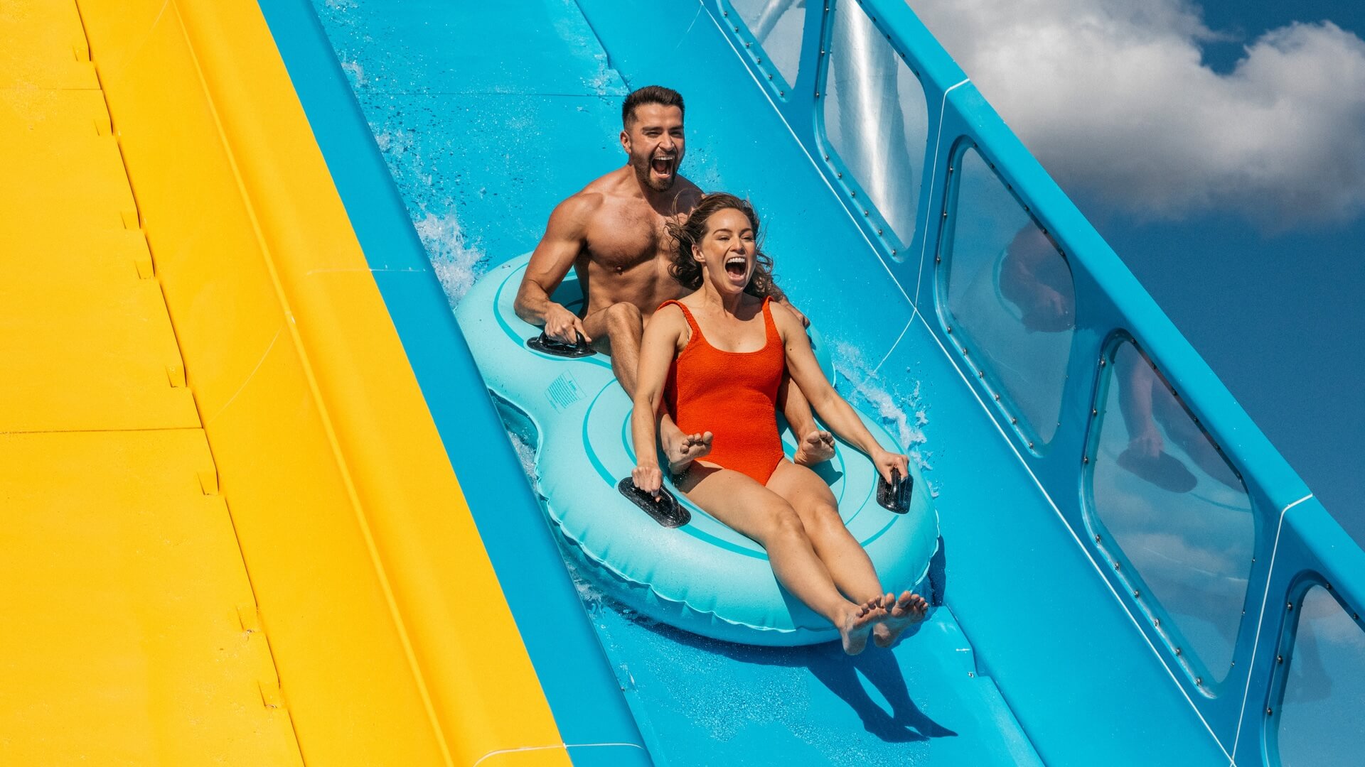 Two people in swimwear riding Kaboom! water slide on a double inflatable tube with blue and yellow sections, appearing excited.