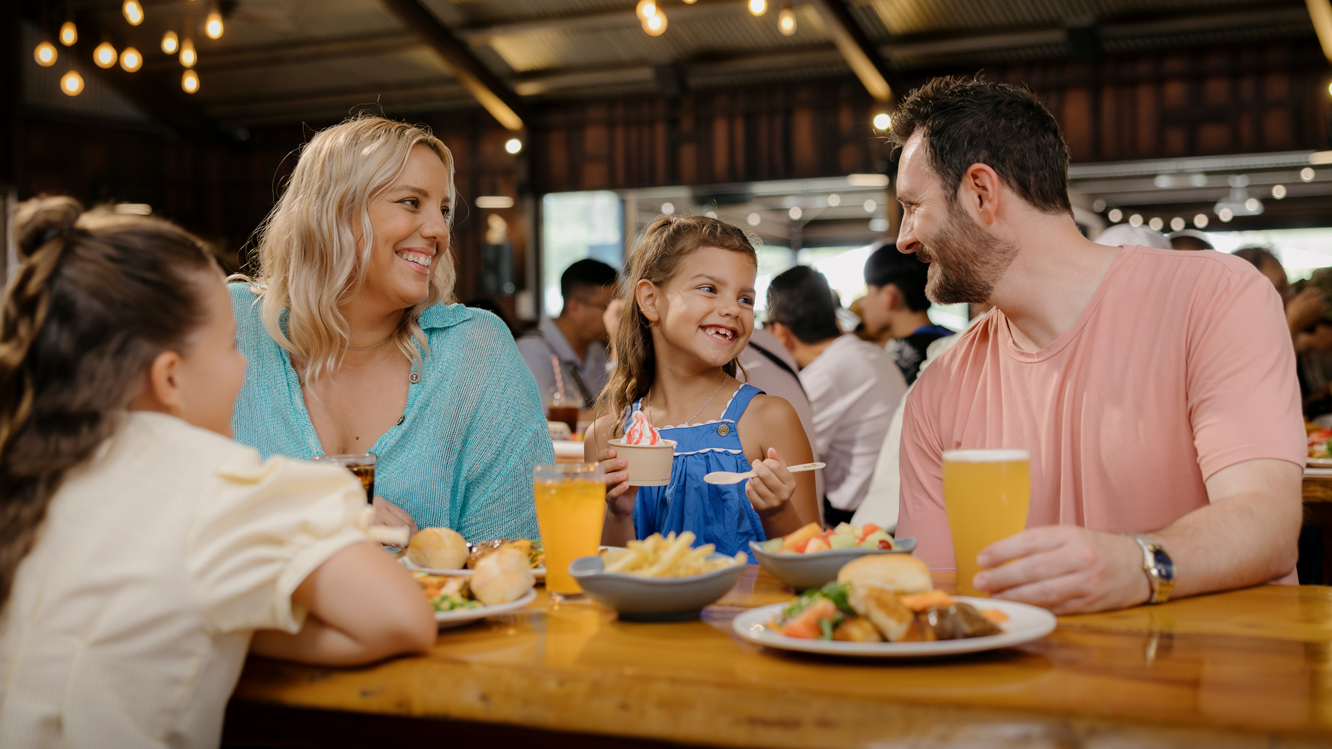 A family of four sits at a restaurant table, smiling and enjoying food and drinks together. Plates of burgers, fries, and fruit are on the table. The setting is warmly lit and lively.
