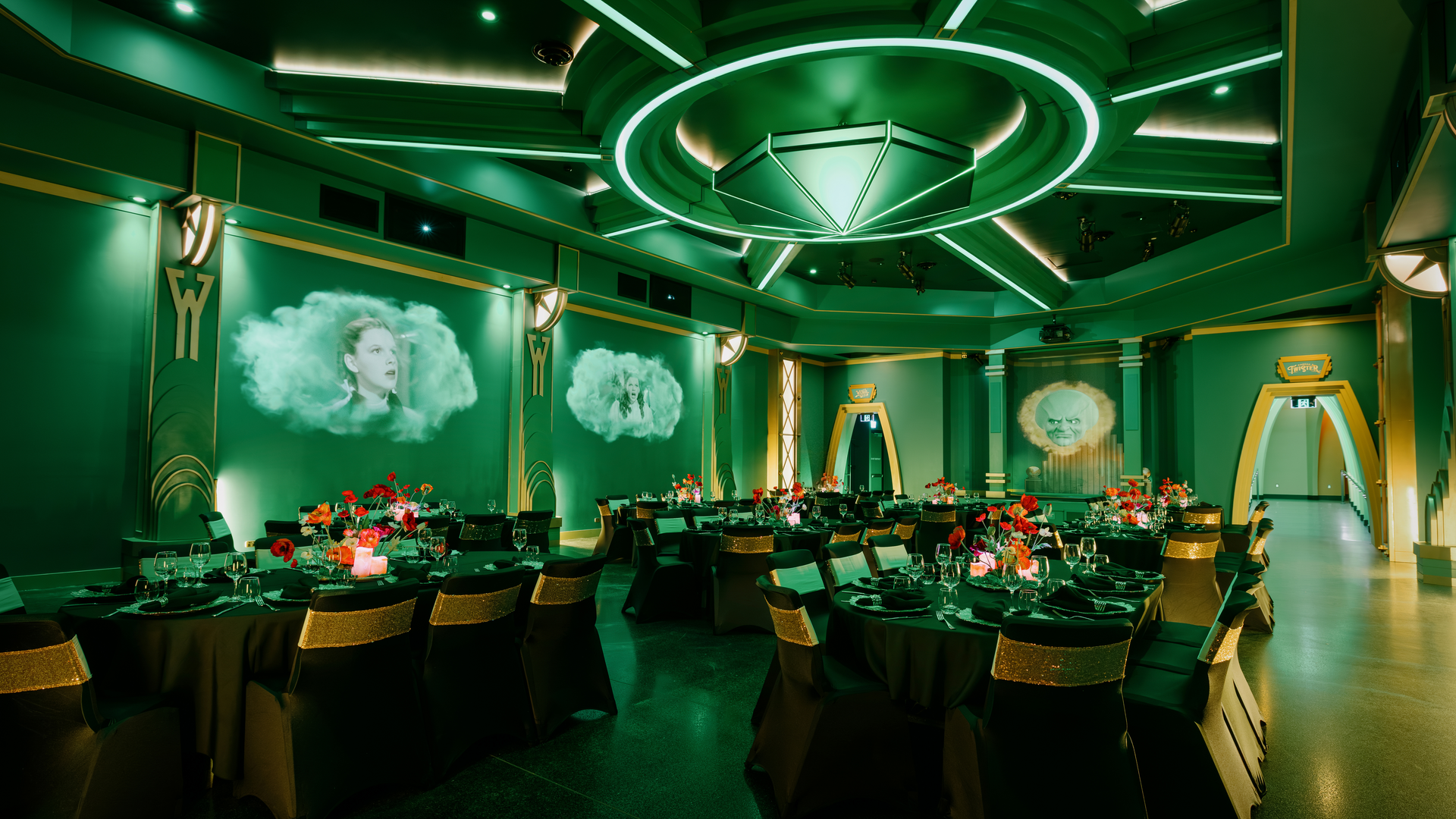 A lavish, green-lit banquet hall with round tables set for an event, red floral centerpieces, black and gold chairs, and large projected images of faces in clouds on the walls. The decor is inspired by "The Wizard of Oz.