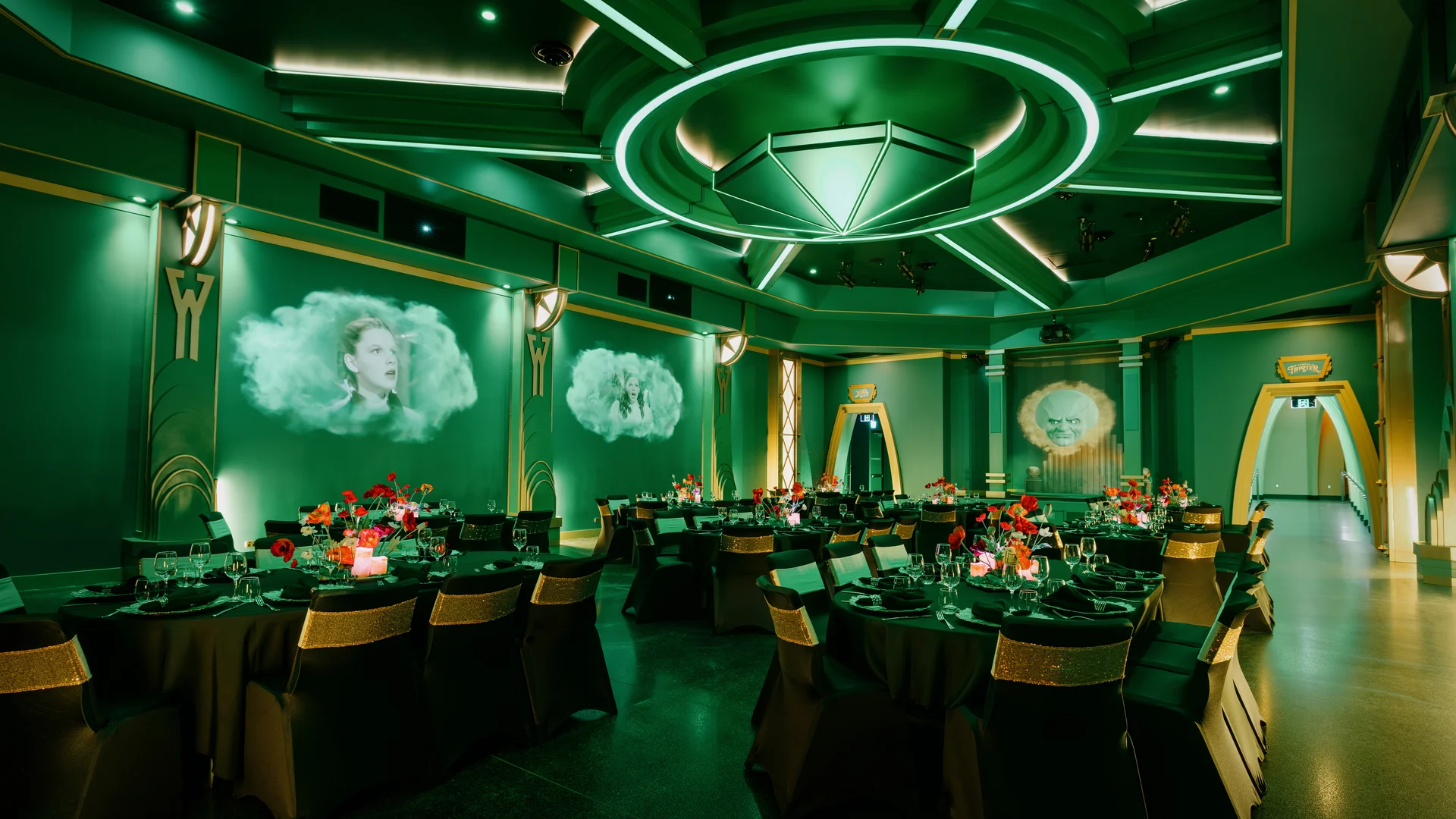 A lavish, green-lit banquet hall with round tables set for an event, red floral centerpieces, black and gold chairs, and large projected images of faces in clouds on the walls. The decor is inspired by "The Wizard of Oz.