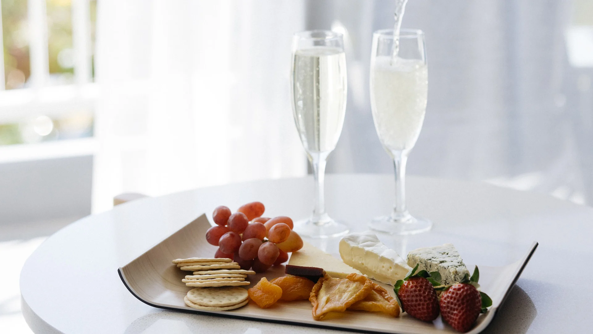 A platter with grapes, crackers, cheese, strawberries, and pastries on a table. Two glasses are being filled with a sparkling beverage in the background.