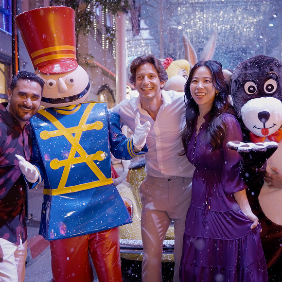 Three adults pose and smile among holiday mascots, including a toy soldier and a bear, under festive lights and falling artificial snow.