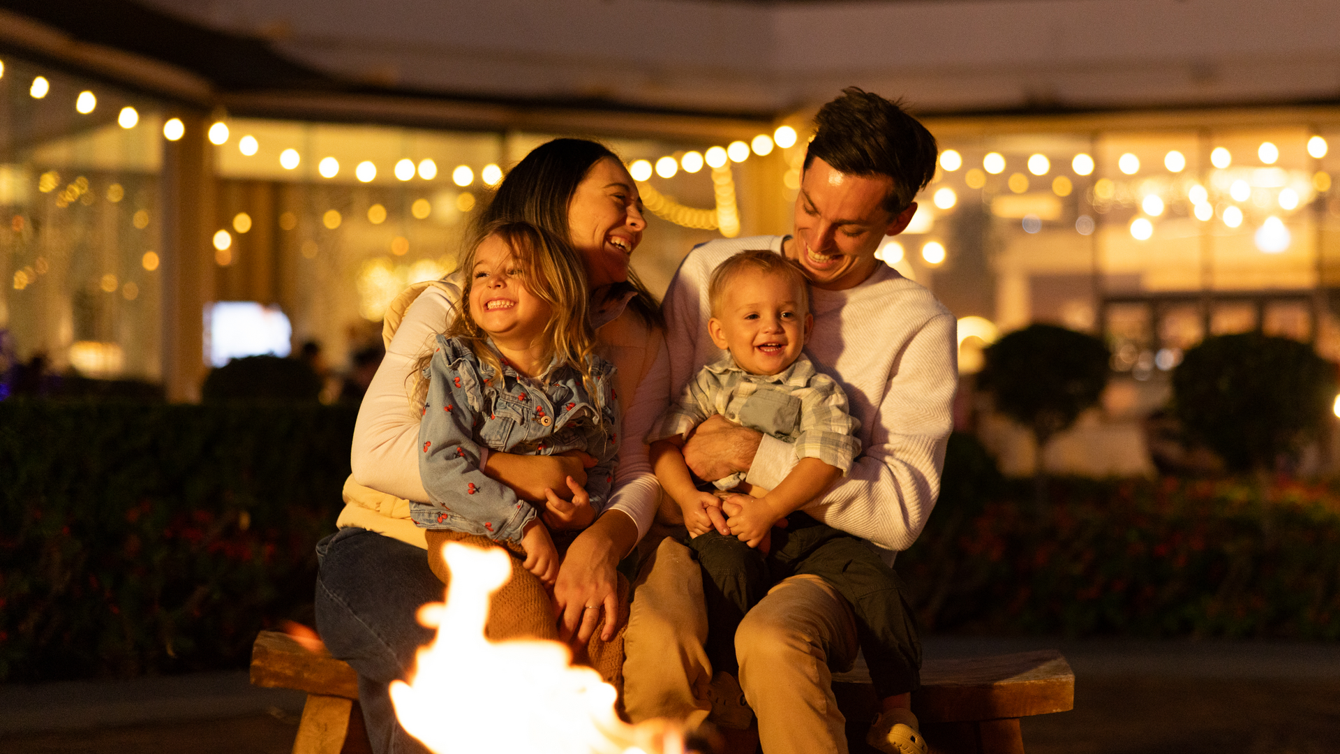 A smiling family of four sits together by an outdoor fire at night, surrounded by warm string lights. The parents hold their two young children, all enjoying the cozy and joyful atmosphere.