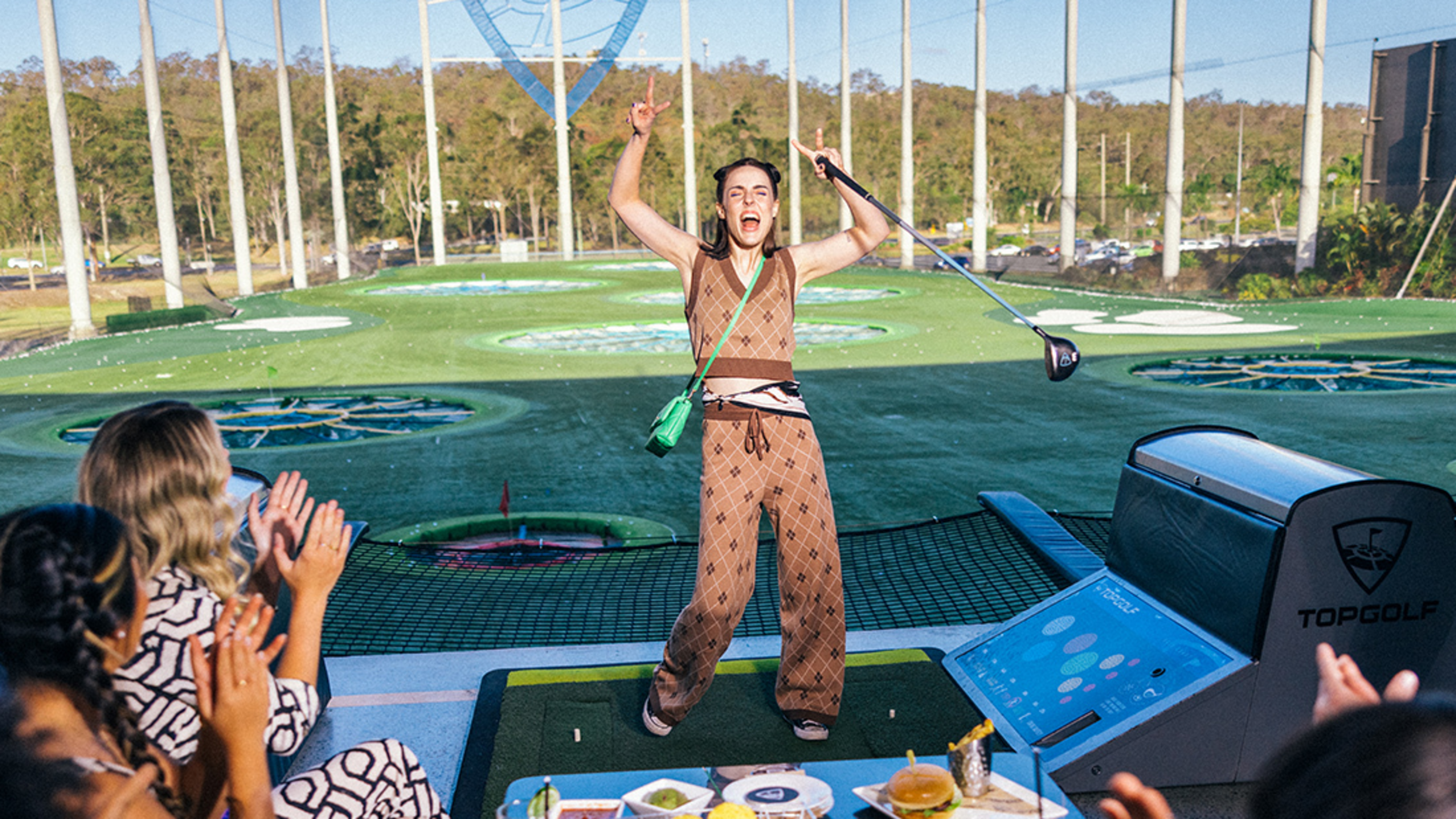 A person celebrates on a golf driving range platform while others applaud. Golf equipment and a monitor are visible.