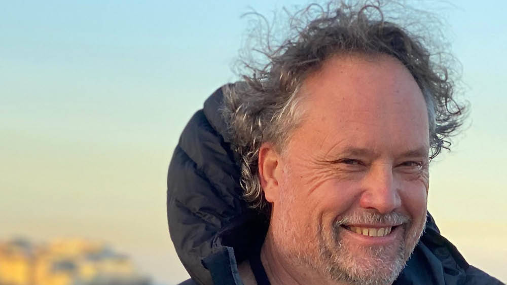 A middle-aged man with curly gray hair and a beard smiles outdoors, wearing a black jacket with a light blue sky in the background.